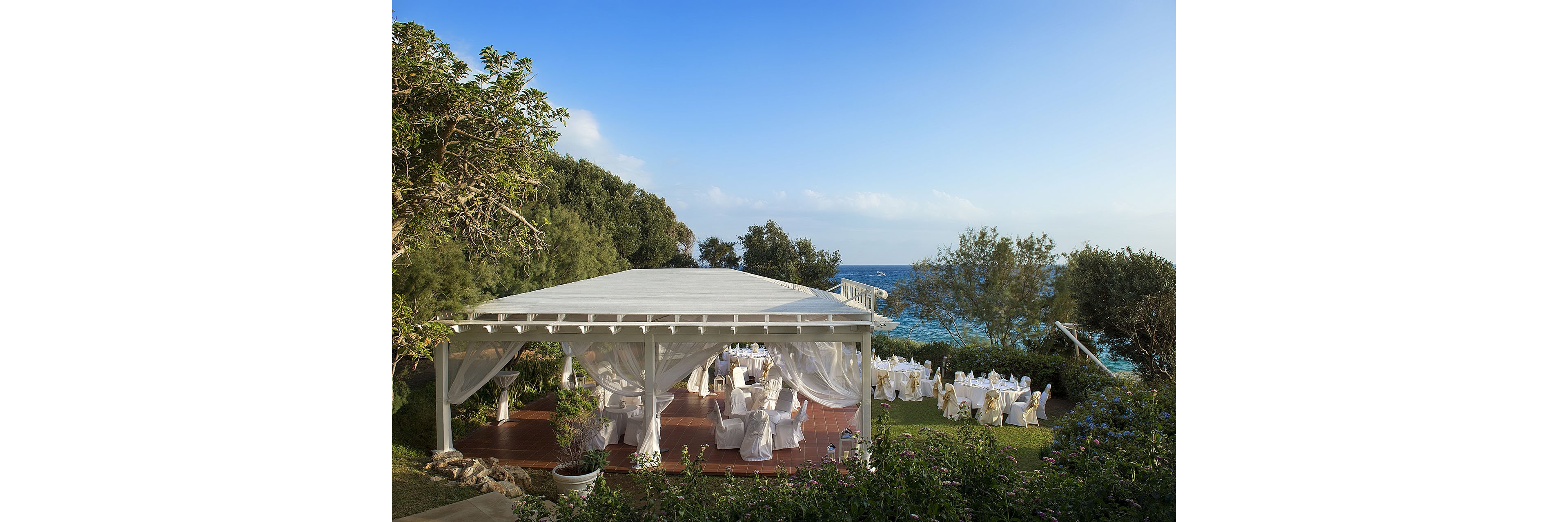 An elegant outdoor wedding reception is set up under a white canopy overlooking the sea. Round tables with white linens and chairs are arranged both under the pavilion and on the surrounding lawn. Lush greenery and trees frame the scene, with blue ocean water visible in the background. The bright daylight and clear sky create a serene and celebratory atmosphere.