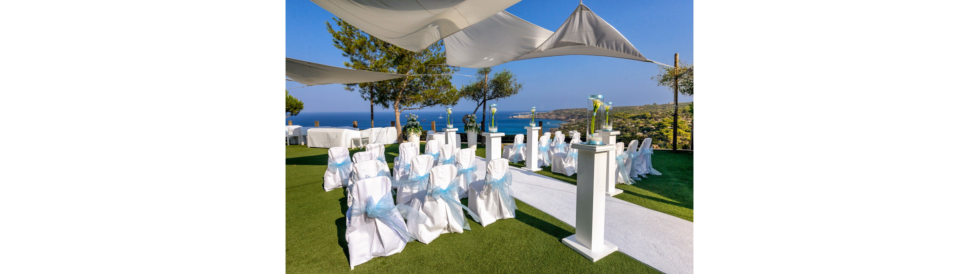 An elegant outdoor wedding ceremony area is arranged on a green lawn overlooking a bright blue sea and coastal landscape. White chairs with light blue sashes are lined along a white aisle runner, facing a simple altar space. Tall white pedestals with glass vases and flowers decorate the aisle, while white fabric canopies provide shade above. Trees and distant hills frame the scenic, sunny setting, creating a fresh and romantic atmosphere.