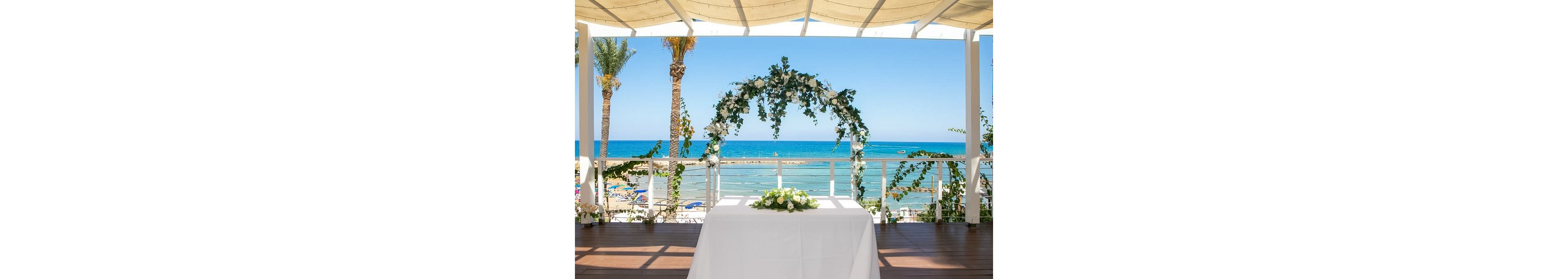 A white-draped altar table is set beneath a floral arch on a terrace overlooking a sunny beach. Greenery and white flowers decorate the arch and railing, framing the turquoise sea and clear blue sky. Palm trees and beach umbrellas are visible in the background, suggesting a coastal resort setting. The scene evokes a romantic, elegant atmosphere prepared for an outdoor wedding ceremony.
