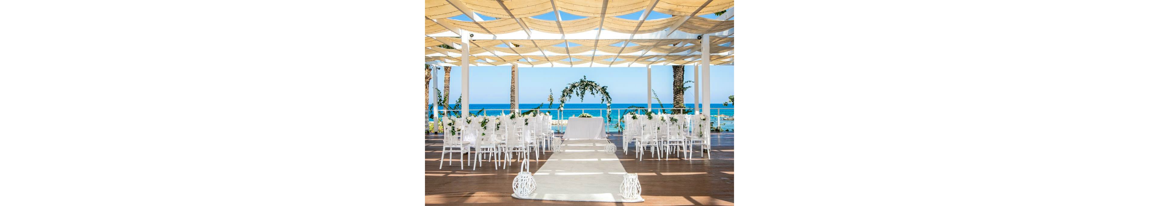 An outdoor wedding ceremony is arranged under a modern pergola overlooking a bright blue sea. White chairs line both sides of a central aisle leading to a table and floral arch decorated with greenery. Lanterns and soft decor elements accent the wooden floor and white runner, creating a fresh, romantic atmosphere. Palm trees and clear skies frame the coastal backdrop, emphasizing a sunny beachside celebration.