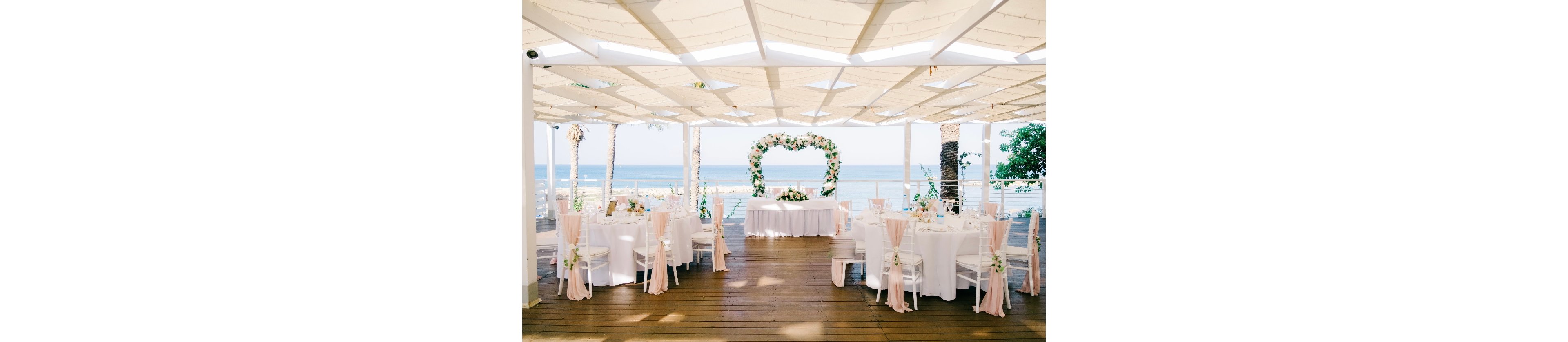 An elegant wedding reception is arranged on a covered seaside terrace overlooking the ocean. Round tables with white linens and soft blush chair sashes surround a central sweetheart table. A large heart-shaped floral arch frames the view of the water, creating a romantic focal point. Soft natural light and pastel flowers contribute to an airy, celebratory atmosphere.