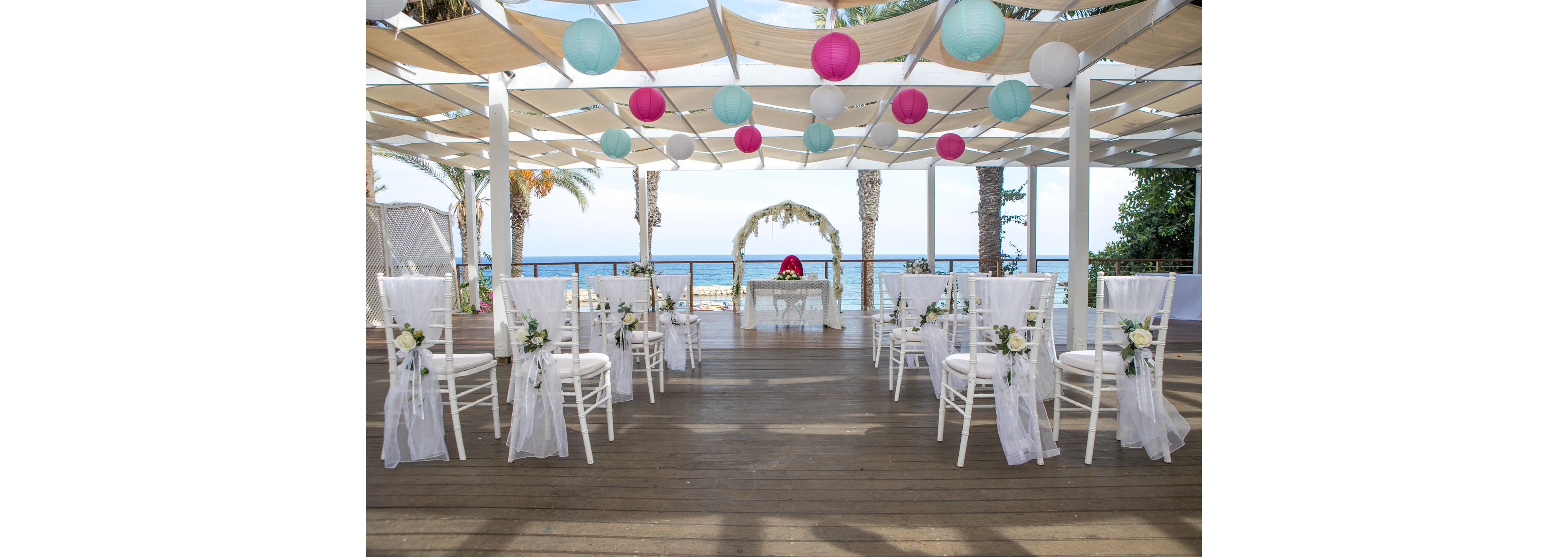 An elegant wedding ceremony area is arranged on a wooden deck overlooking the sea. White chairs decorated with flowers and sheer fabric form two rows facing a floral arch and table at the front. Colorful paper lanterns in white, pink, and turquoise hang from a canopy structure above. Palm trees and a clear blue ocean provide a bright, summery backdrop, creating a romantic coastal atmosphere.