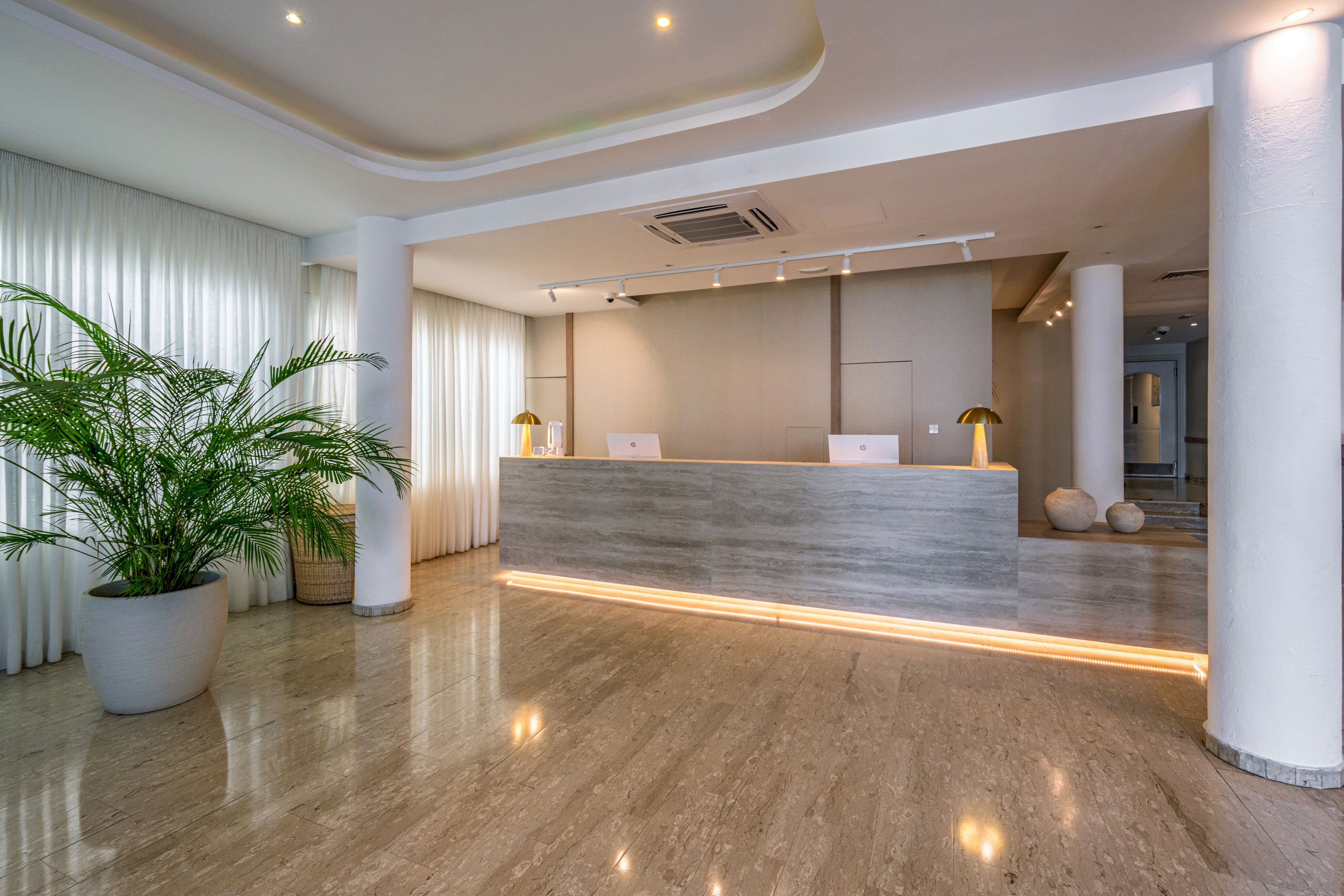A contemporary reception desk stands in a spacious hotel lobby with a polished marble floor. Soft ceiling lighting and an illuminated base under the counter create a warm, welcoming atmosphere. Large white curtains line the wall behind a tall potted plant, adding texture and softness to the space. Minimalist decor and neutral colors emphasize a clean, modern interior design.