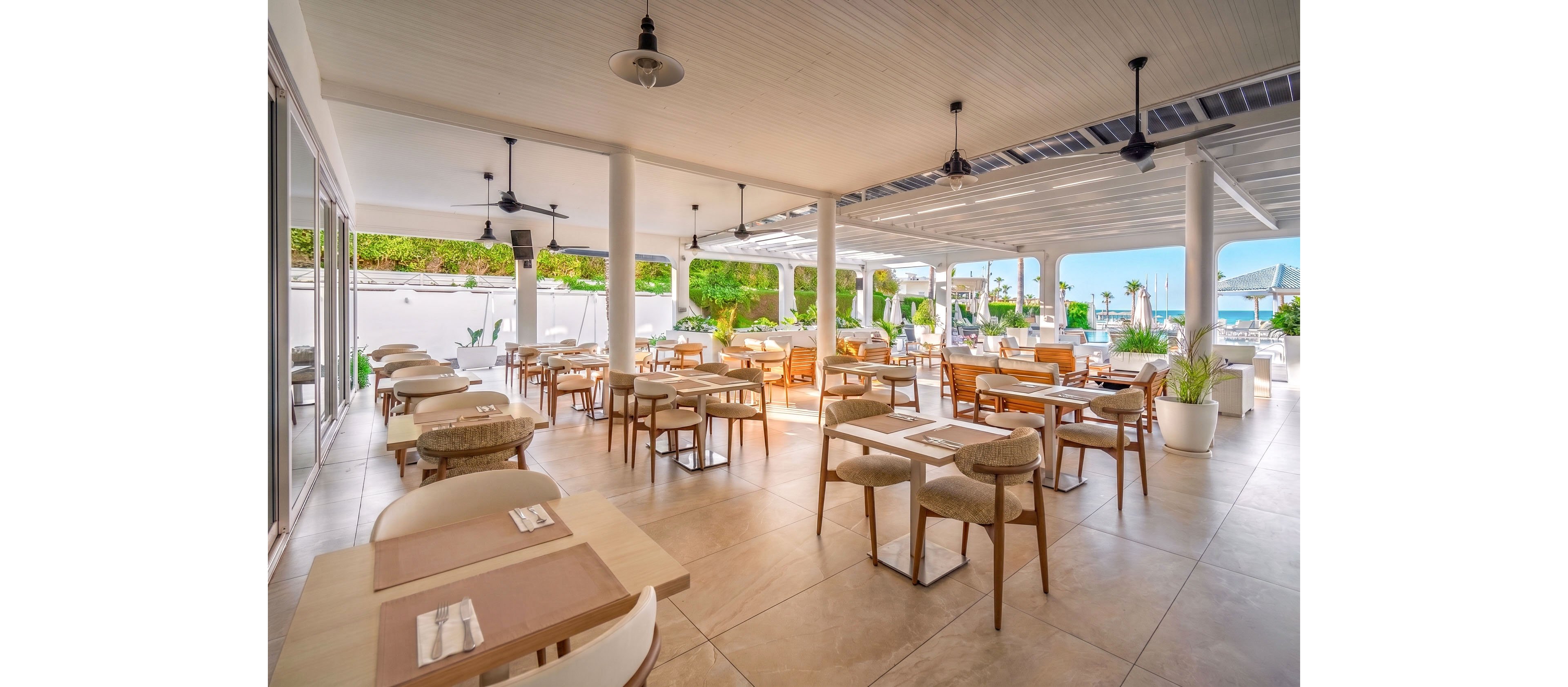 Bright open-air restaurant features neatly arranged wooden tables and cushioned chairs under a white covered terrace. Large openings reveal a view of lush greenery and the distant blue sea, creating a relaxed coastal atmosphere. Neutral beige flooring and light-toned furniture contribute to a clean, modern aesthetic. Table settings are minimal, suggesting a casual yet refined dining experience.