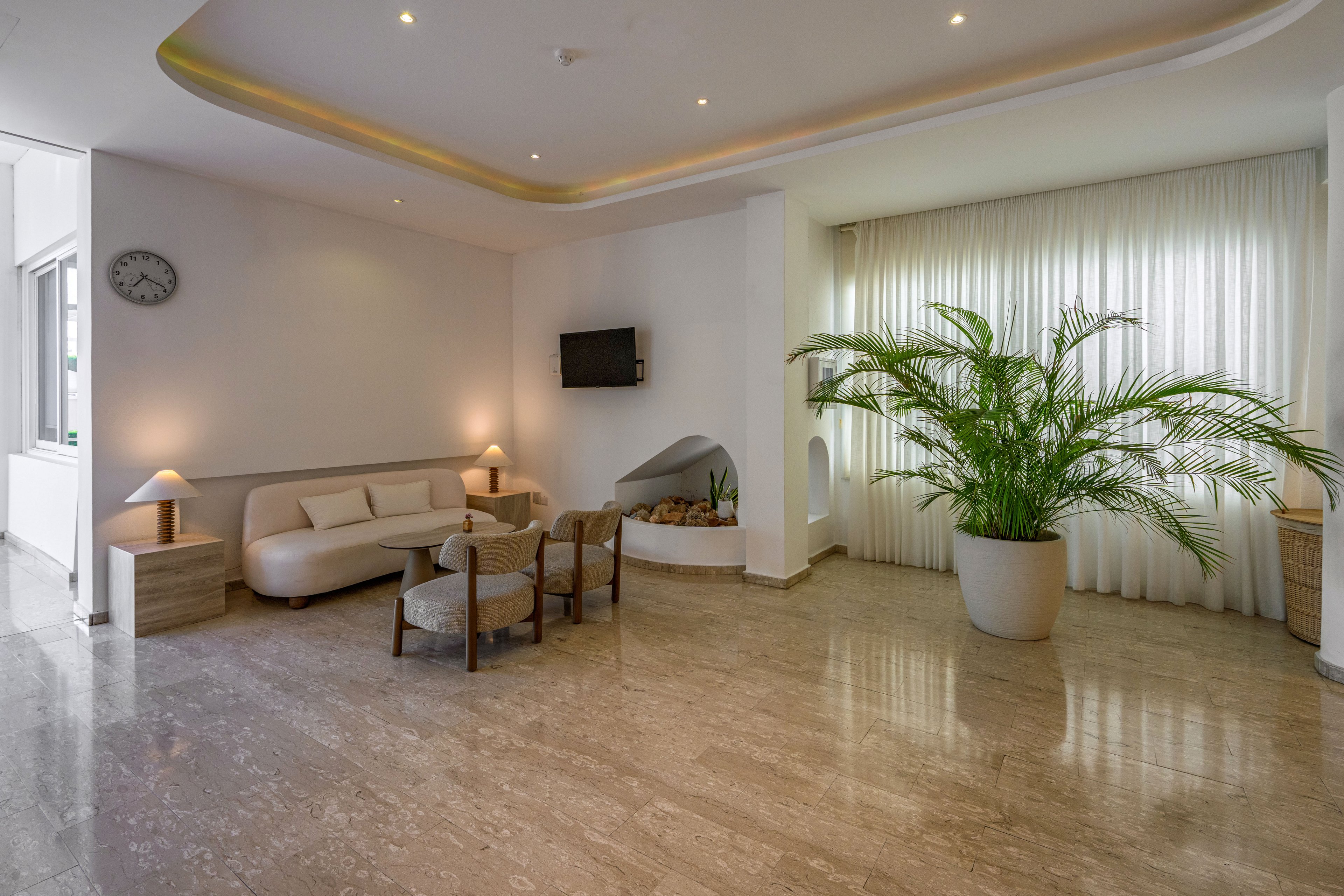 A contemporary lobby lounge features a light-colored sofa and small chairs arranged around a low table. The space is illuminated by recessed ceiling lights and two table lamps, creating a warm and inviting atmosphere. A wall-mounted television and a round wall clock add functional details to the minimalist decor. Large sheer curtains diffuse daylight behind a tall potted plant, enhancing the airy, spacious feel of the room.