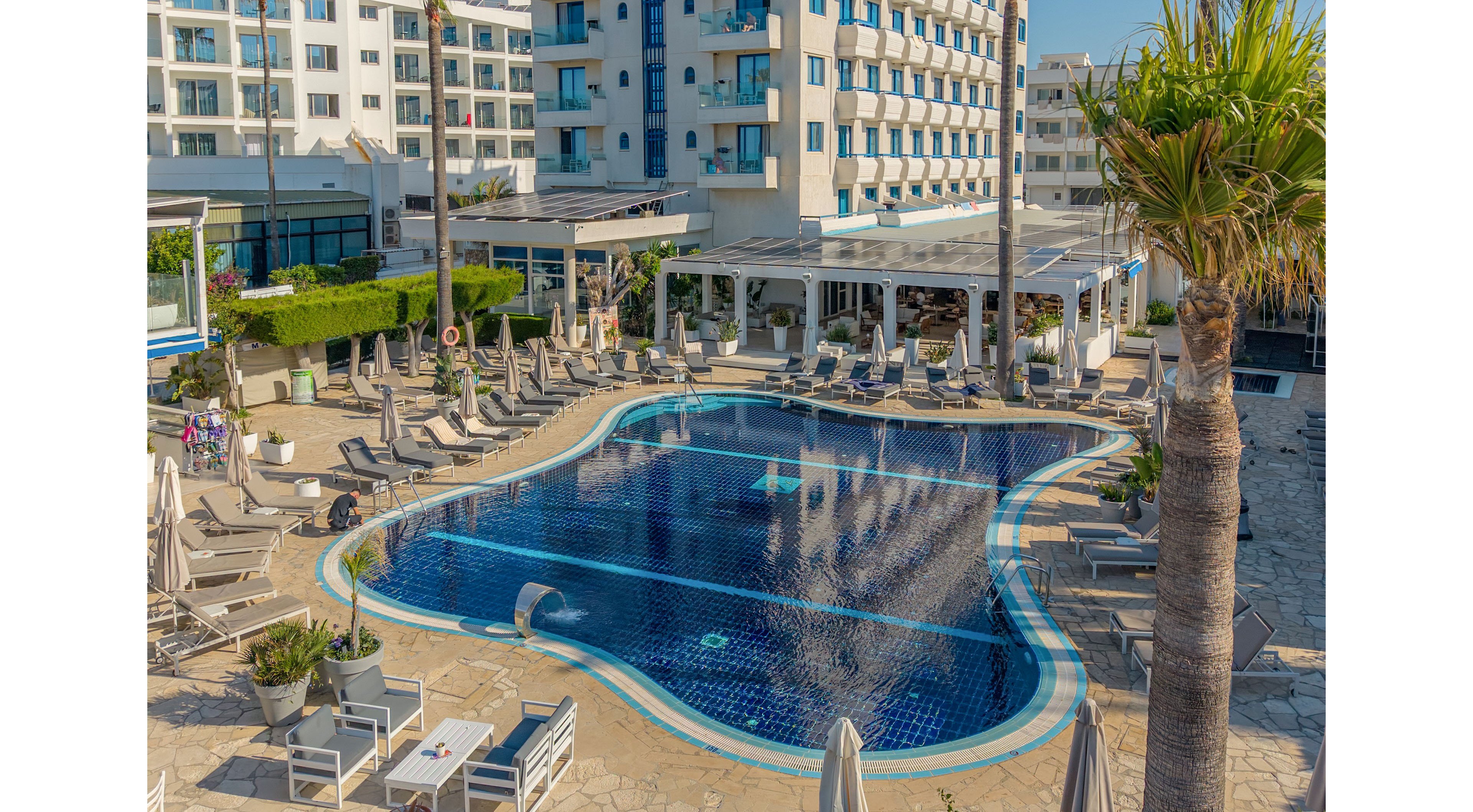 An outdoor hotel swimming pool sits at the center of a sunlit courtyard surrounded by lounge chairs and umbrellas. Multi-story white buildings with balconies rise in the background, suggesting a resort or vacation setting. Palm trees and neatly trimmed greenery add a tropical touch to the scene. The clear blue water and tiled pool surface create a relaxing atmosphere under bright daylight.