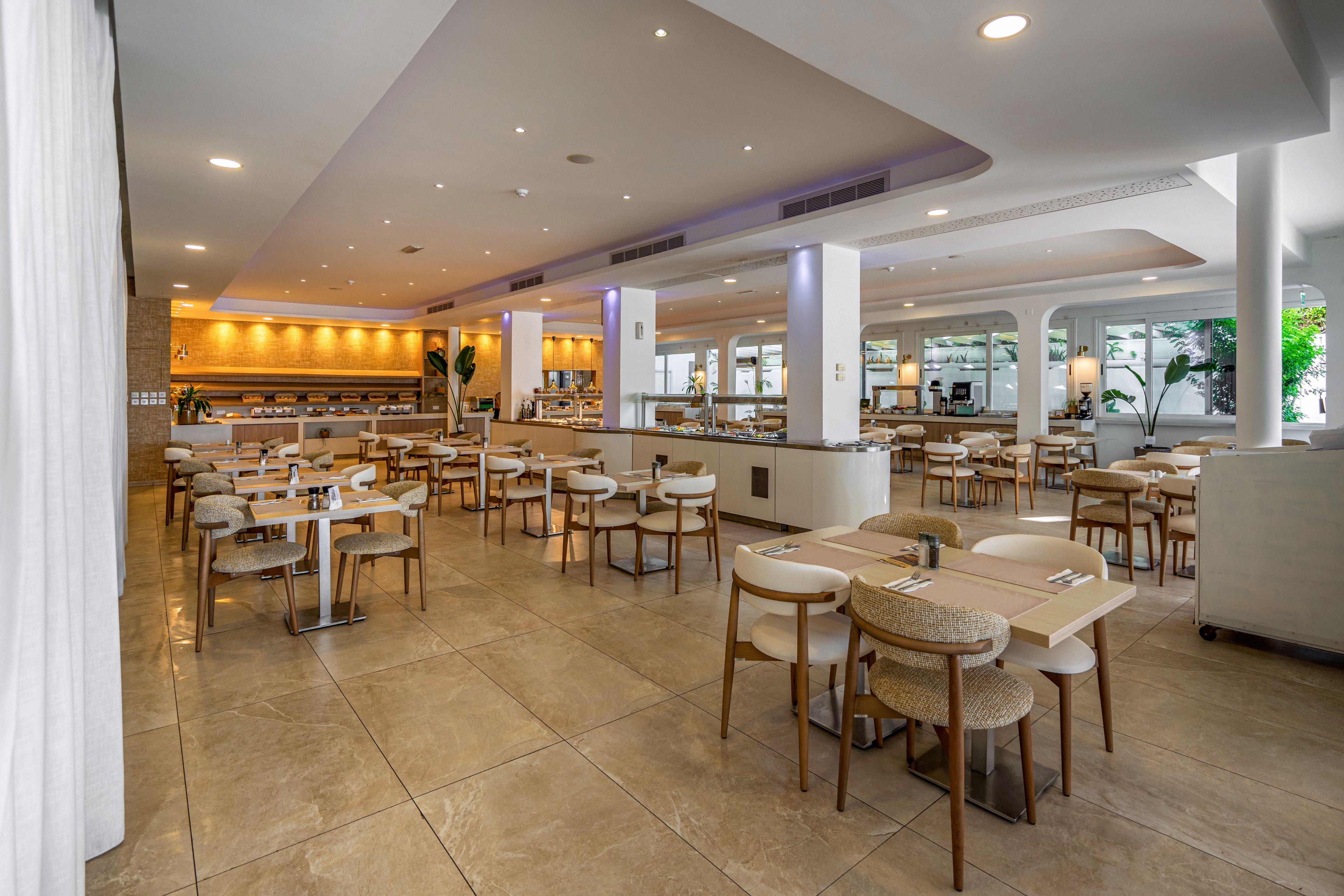Spacious contemporary restaurant dining area with neatly arranged tables and chairs. The room features large floor tiles, soft neutral colors, and recessed ceiling lighting. Wooden furniture and light upholstery create a warm, minimalist aesthetic. Floor-to-ceiling windows and open archways bring in natural light and views of greenery.