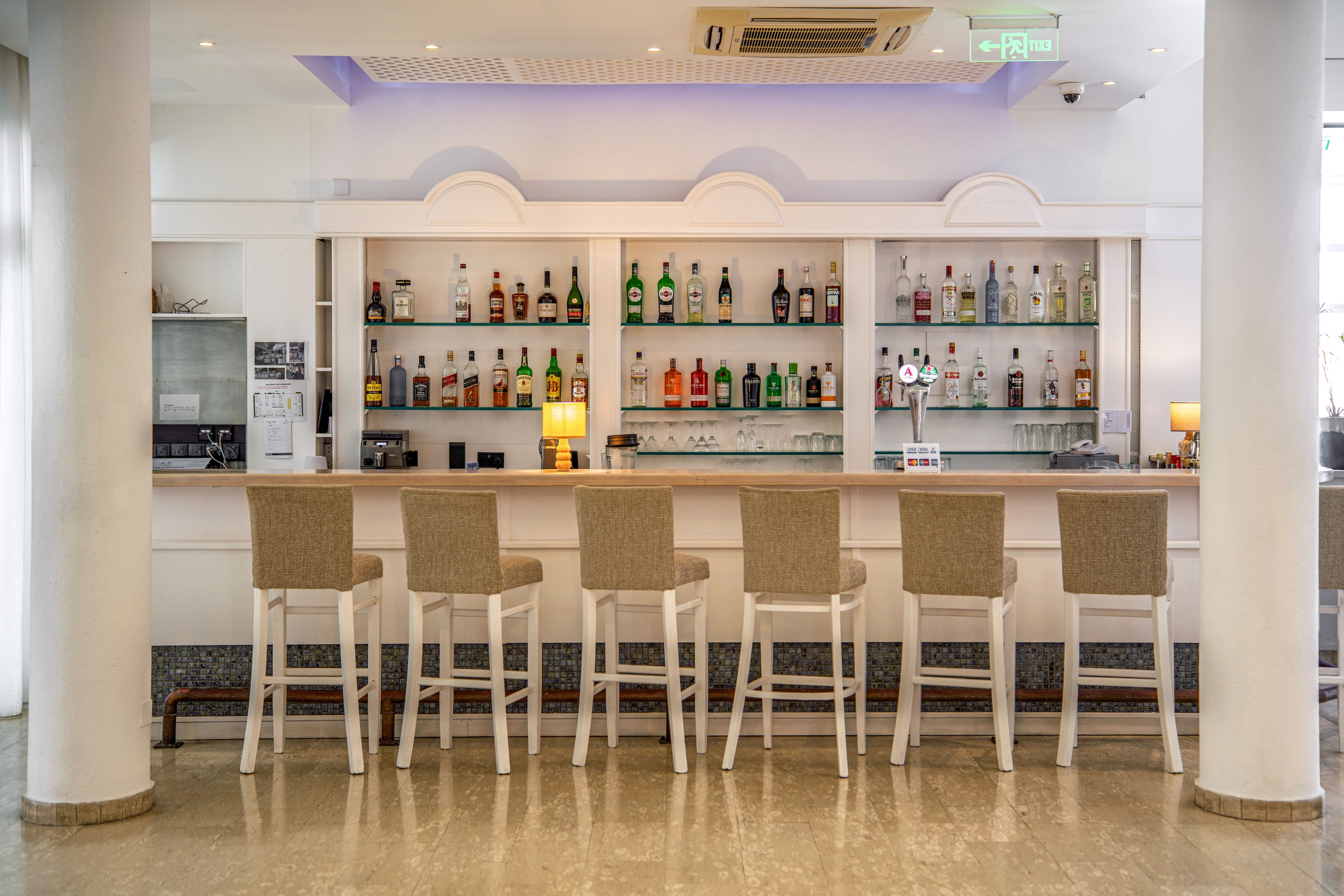 A contemporary bar interior features a long white counter with six beige upholstered bar stools neatly aligned. Behind the counter, shelves display an assortment of liquor bottles and glassware, creating a colorful focal point. The space is bright and clean, with white columns, polished flooring, and soft ceiling lighting that adds a relaxed atmosphere. No people are present, emphasizing the tidy, ready-for-service setting.