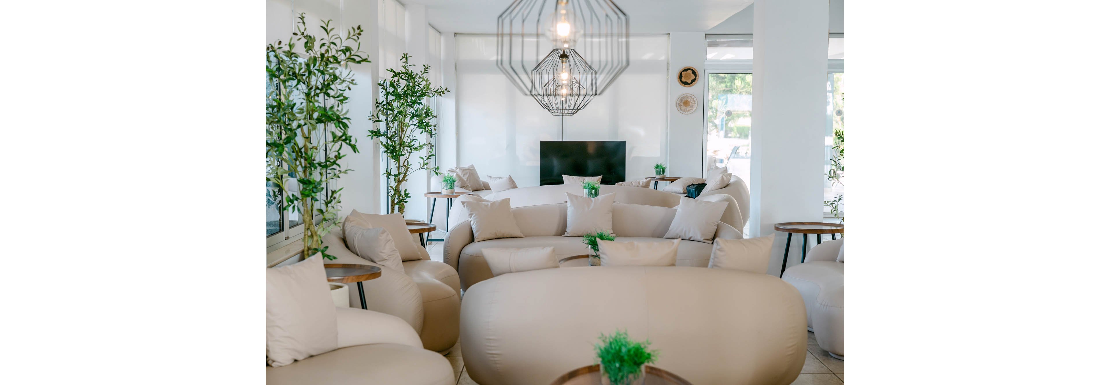 A bright contemporary lounge features large beige inflatable sofas and armchairs arranged in a cozy layout. Tall green plants in pots and small round wooden side tables add natural warmth to the minimalist white interior. Geometric pendant lights hang from the ceiling, illuminating the space with soft light. A flat-screen television is positioned against the far wall, completing the relaxed social setting.