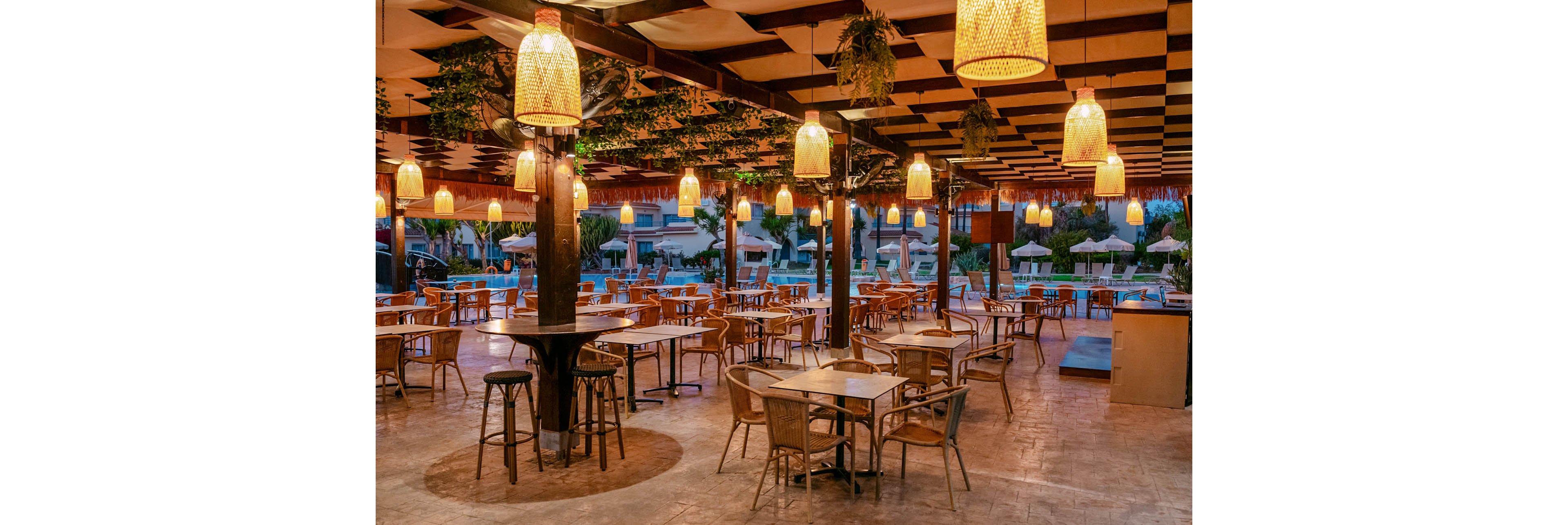 An open-air restaurant terrace is shown at dusk with numerous empty wooden tables and wicker chairs neatly arranged. Warm hanging lanterns illuminate the covered patio, creating a cozy and inviting atmosphere. In the background, a swimming pool area with sun loungers and umbrellas is visible, suggesting a resort or hotel setting. The overall scene feels relaxed and ready for guests to arrive for evening dining.