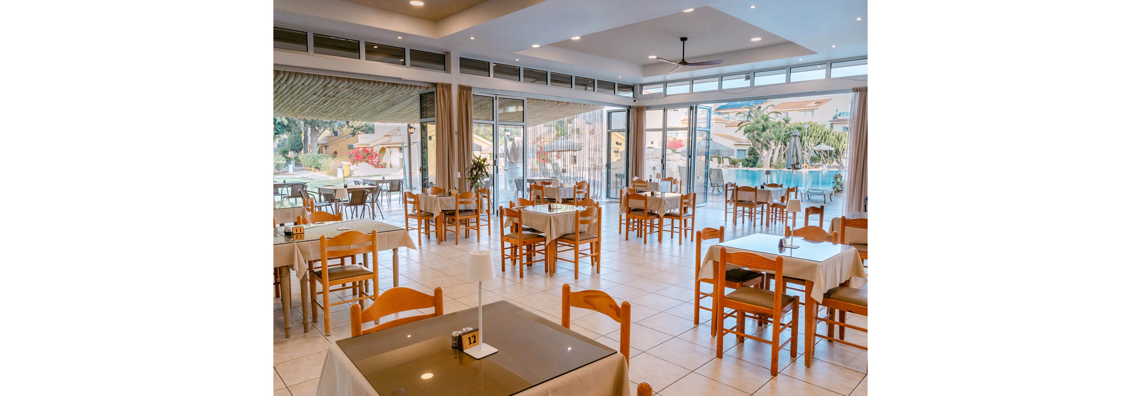 Spacious restaurant dining area with wooden chairs and neatly set tables stands empty. Large floor-to-ceiling windows open to an outdoor patio and poolside view with palm trees and buildings. Neutral tile flooring and light-colored tablecloths create a clean, airy atmosphere. A small table sign with the number 12 is visible in the foreground.