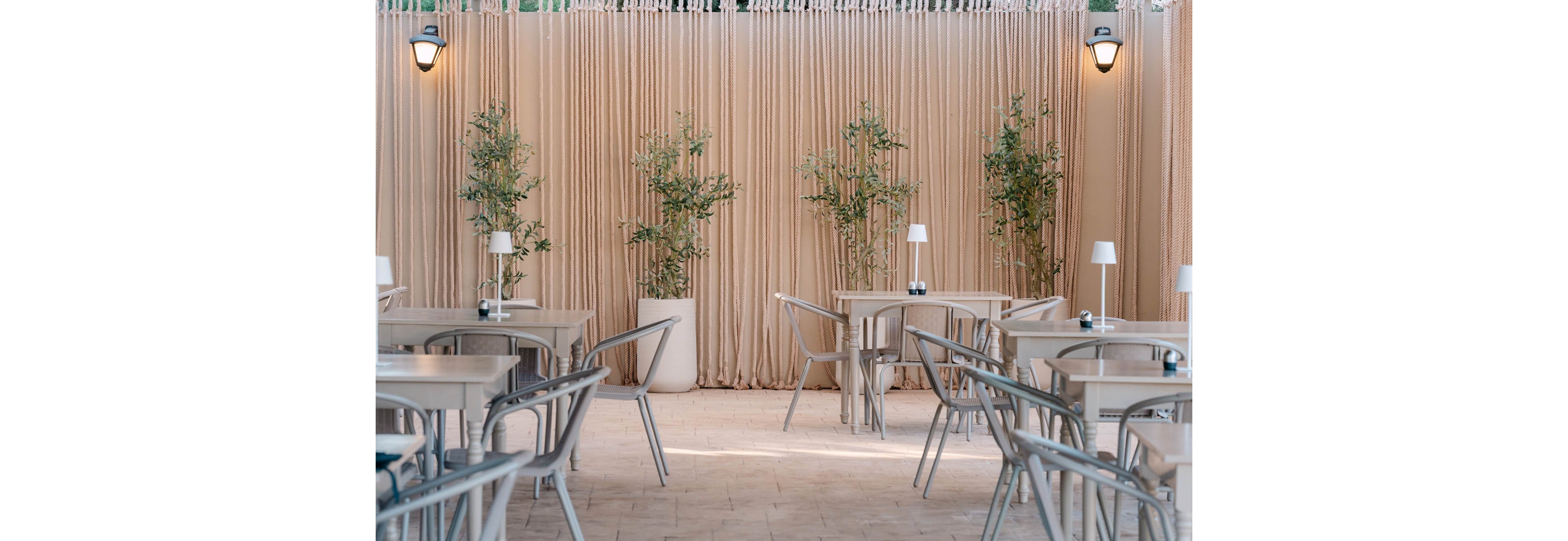 An outdoor cafe features light-colored tables and metal chairs arranged neatly on a tiled floor. Small table lamps and condiment holders sit on the tabletops, adding a modern touch. Tall potted plants are placed against a backdrop of beige curtains, creating a soft, elegant atmosphere. Wall-mounted lights above the curtains contribute to the cozy, inviting setting.