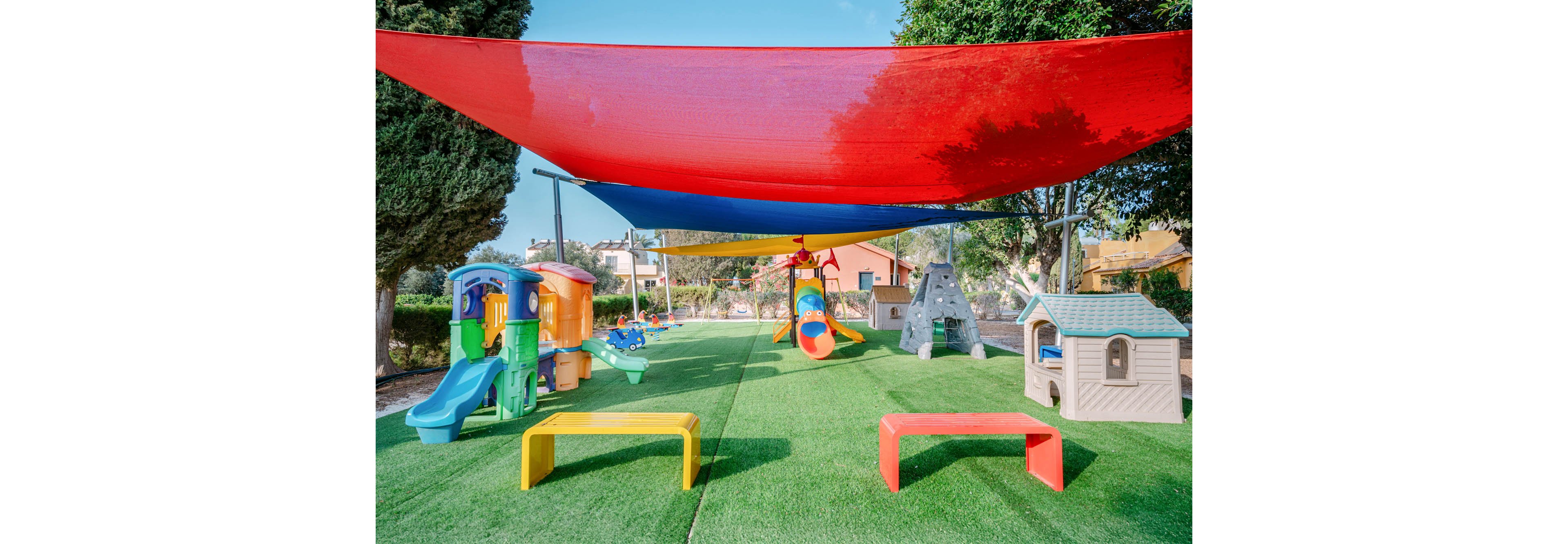 A bright outdoor playground features multiple plastic play structures and slides on a green artificial turf surface. Red and blue fabric canopies stretch overhead, providing shade across the play area. A small playhouse, climbing wall, and airplane-themed rocker are arranged around the space. Trees and nearby houses frame the background, suggesting a residential or community setting.