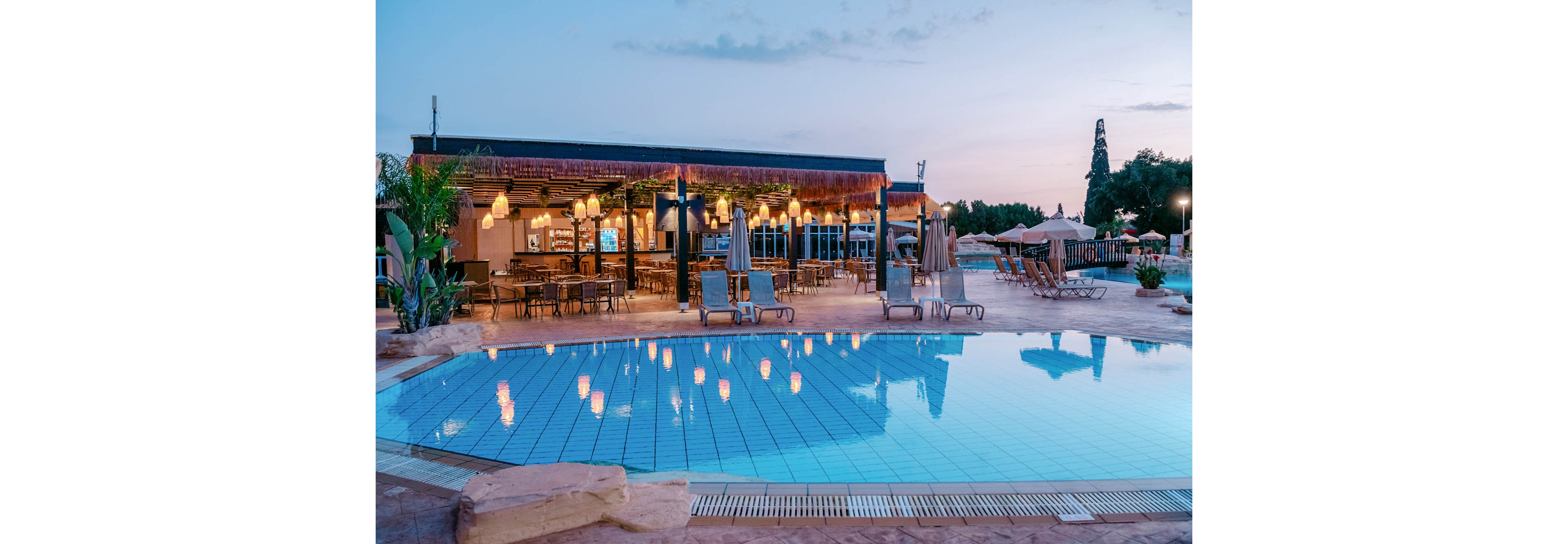 An illuminated outdoor swimming pool reflects the warm lights of a nearby resort bar at dusk. Lounge chairs and closed umbrellas line the poolside terrace, creating a relaxed vacation atmosphere. The bar area features hanging lights, tables, and seating under a covered structure. Soft evening sky tones and surrounding greenery enhance the tranquil, holiday setting.