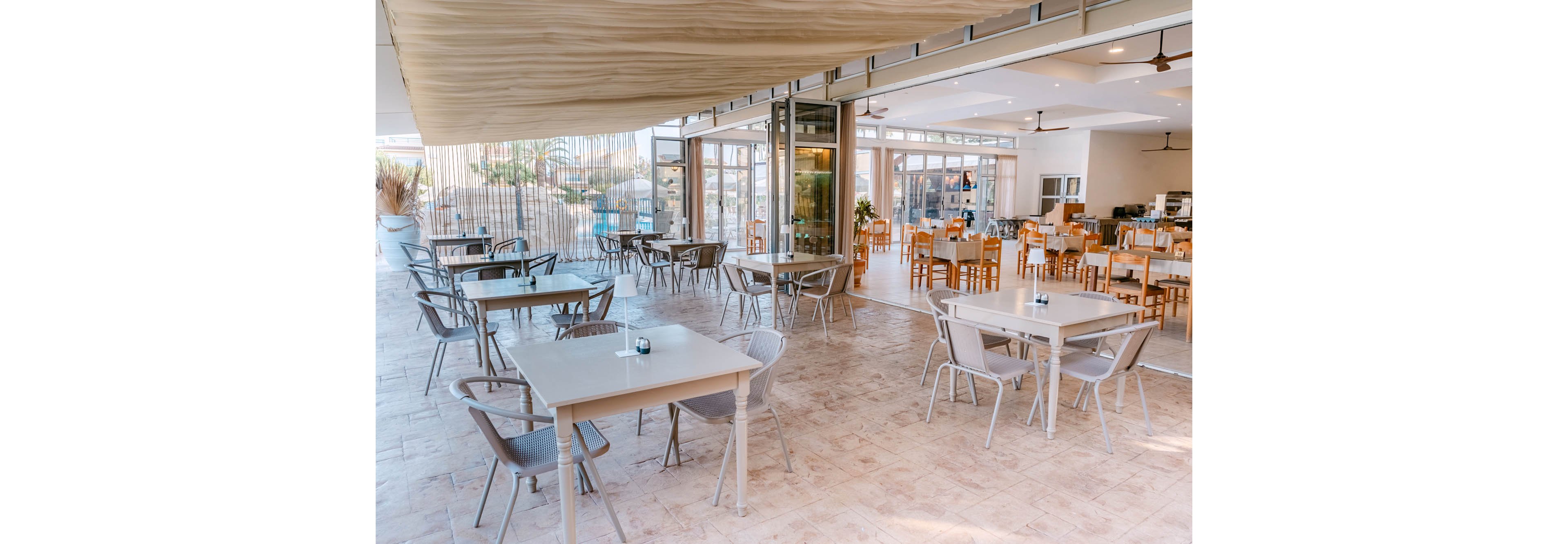 A contemporary open-air restaurant features neatly arranged empty tables and chairs. The space opens onto an indoor dining area through large sliding glass doors. Neutral tones, light wood furniture, and tiled flooring create a clean, airy atmosphere. Ceiling fans and natural light enhance the relaxed, coastal-inspired setting.
