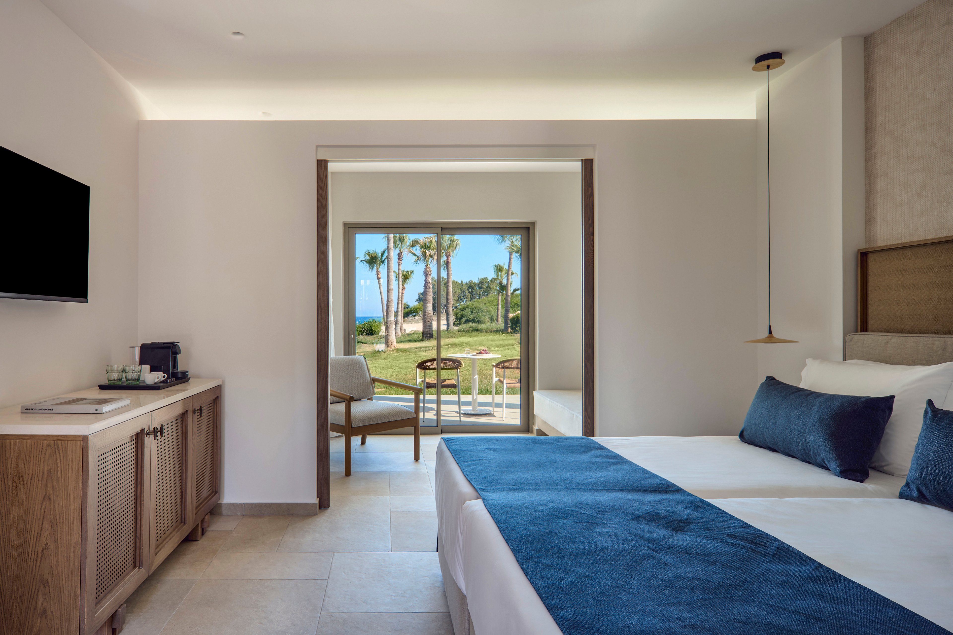 Contemporary hotel room features twin beds with white linens and blue accent throws. A large sliding glass door opens to a patio with chairs and a view of palm trees and green lawns. Minimalist furnishings include a wall-mounted television, wooden sideboard, and a small coffee station. Soft natural light and neutral tones create a calm, relaxing atmosphere ideal for vacation or leisure stays.