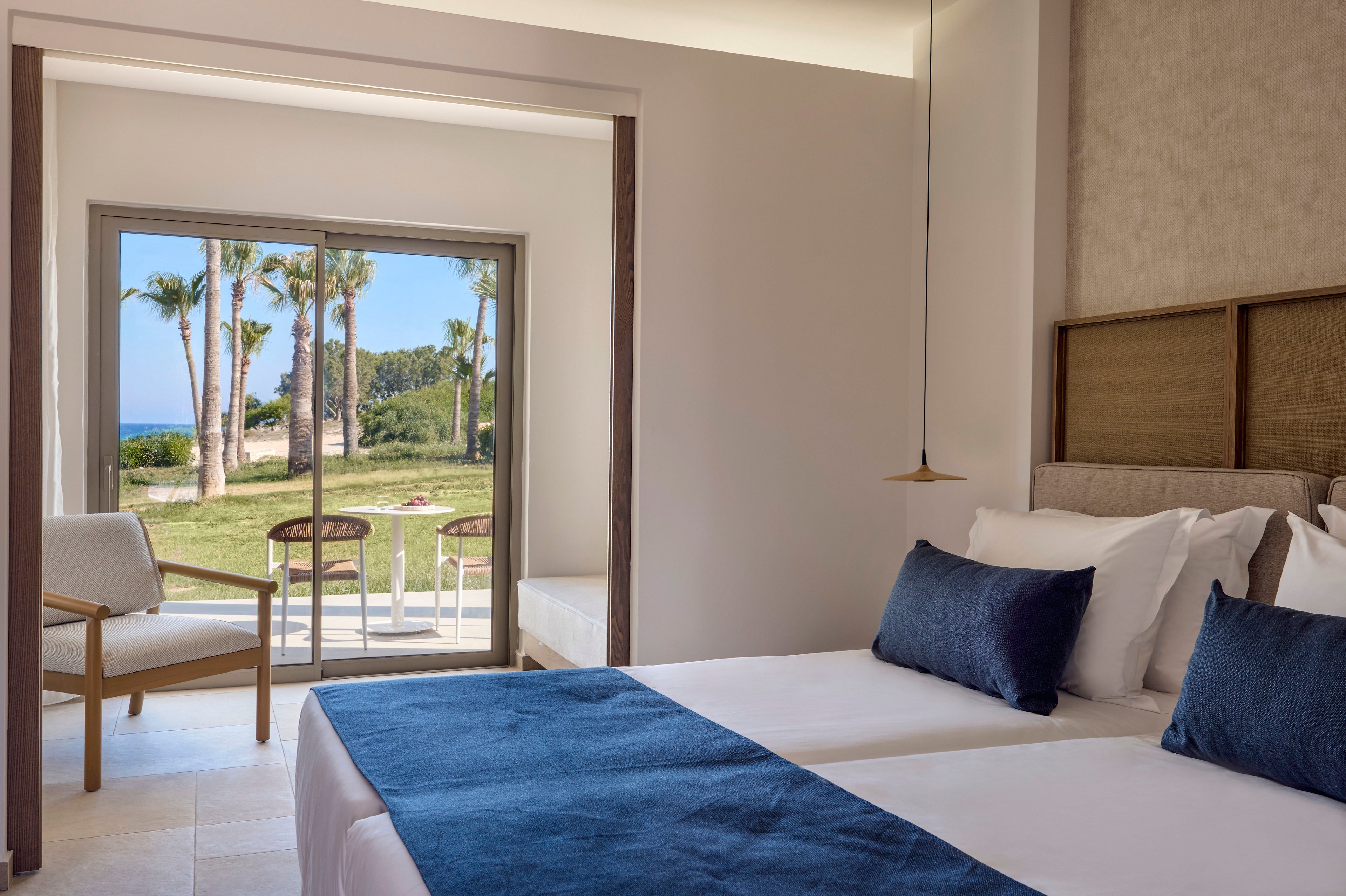 Bright contemporary hotel room features two twin beds with white linens and blue accent pillows. A large sliding glass door opens to a patio with chairs, overlooking a grassy garden and palm trees. Neutral walls and minimalist furniture create a clean, relaxing atmosphere. In the distance, the blue sea is visible beyond the landscaped grounds.
