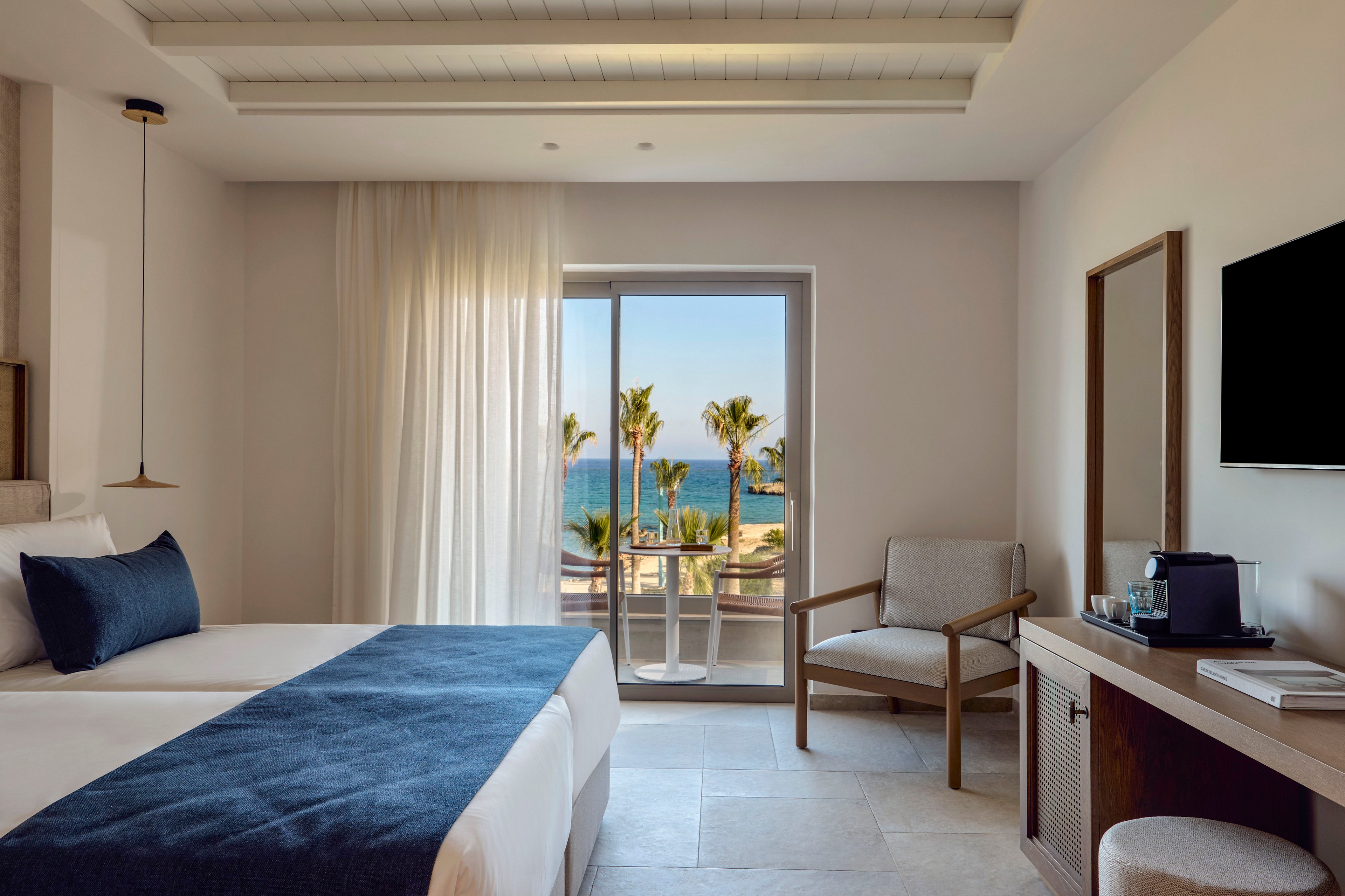 Contemporary hotel room interior featuring twin beds with white linens and blue accents. A sliding glass door opens to a balcony overlooking palm trees and the ocean. The space includes a lounge chair, desk area with coffee machine, and wall-mounted television. Soft natural light filters through sheer curtains, creating a calm and relaxing atmosphere.