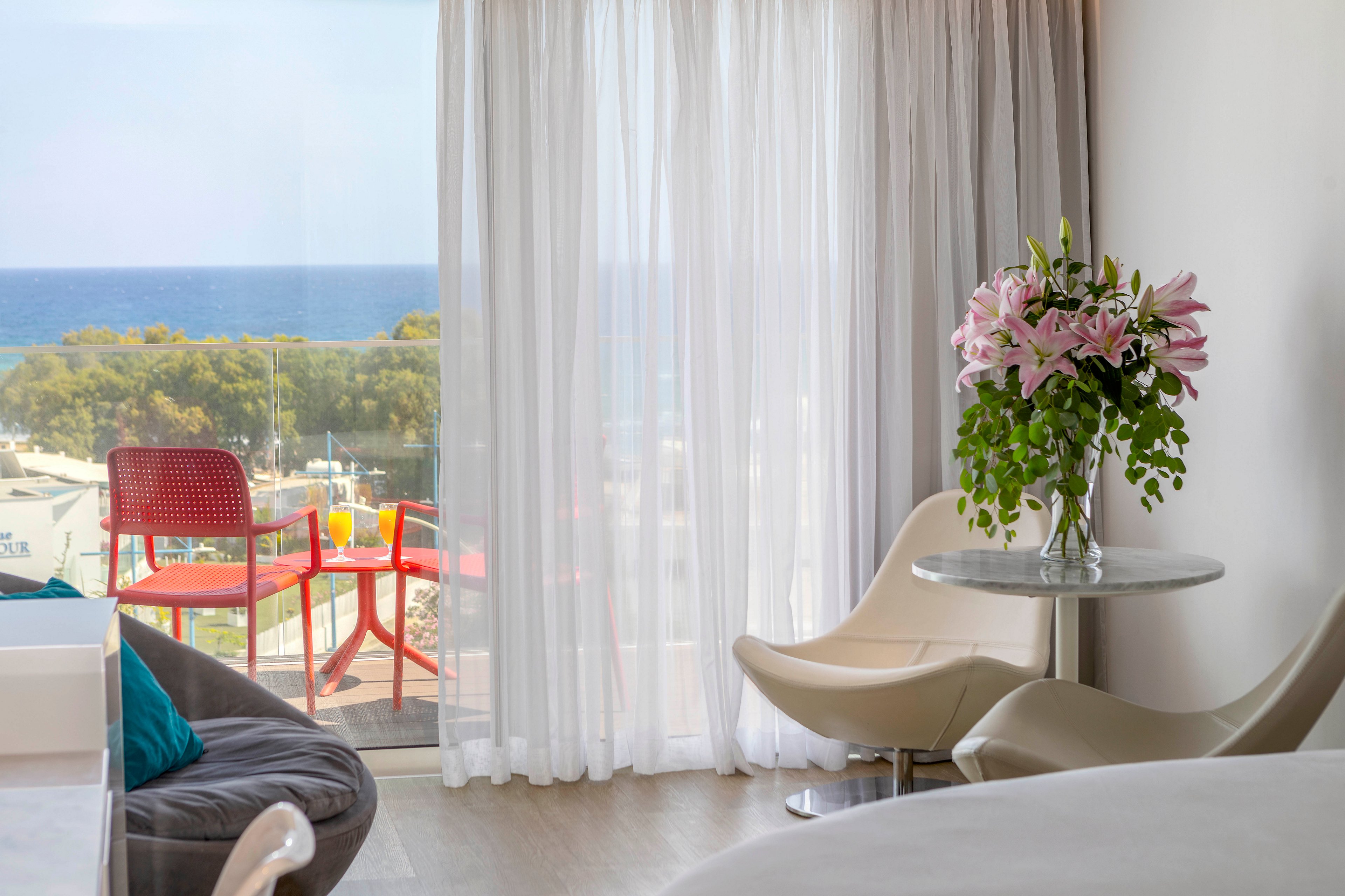 A modern, sunlit hotel room features contemporary chairs and a small round table with a bouquet of pink lilies. Sheer white curtains frame a glass door leading to a balcony overlooking the sea and distant trees. On the balcony, two red chairs and a small table with drinks add a vibrant accent to the serene coastal setting. Soft neutral tones inside contrast with the bright colors outside, creating a relaxed vacation atmosphere.