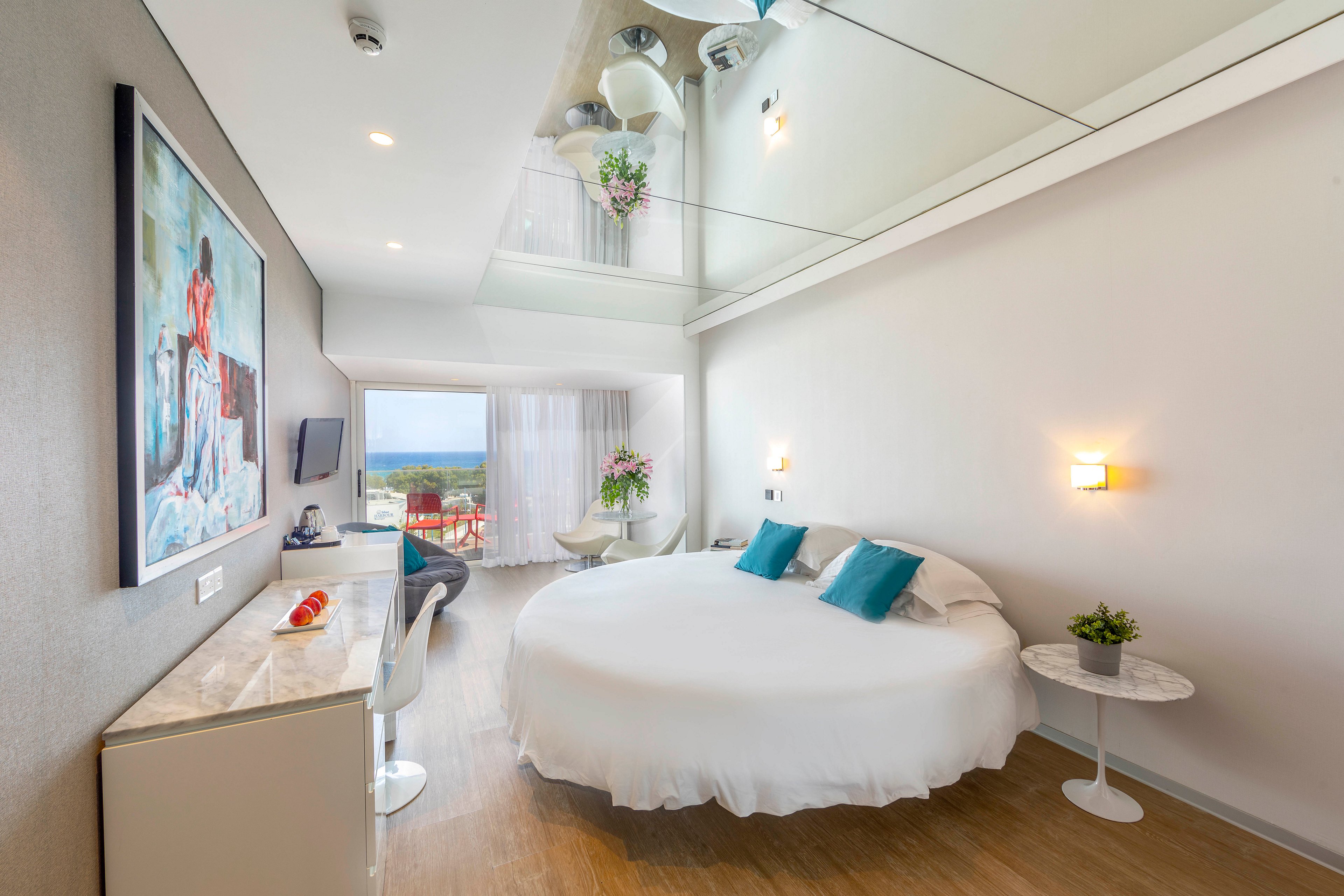 Bright contemporary hotel room featuring a large round bed with teal accent pillows. The space includes a sleek desk area, wall-mounted television, and artwork on the wall. Floor-to-ceiling windows open to a balcony with outdoor seating and a view of the landscape. Light wood flooring, white furniture, and mirrored ceiling elements create an airy, spacious feel.