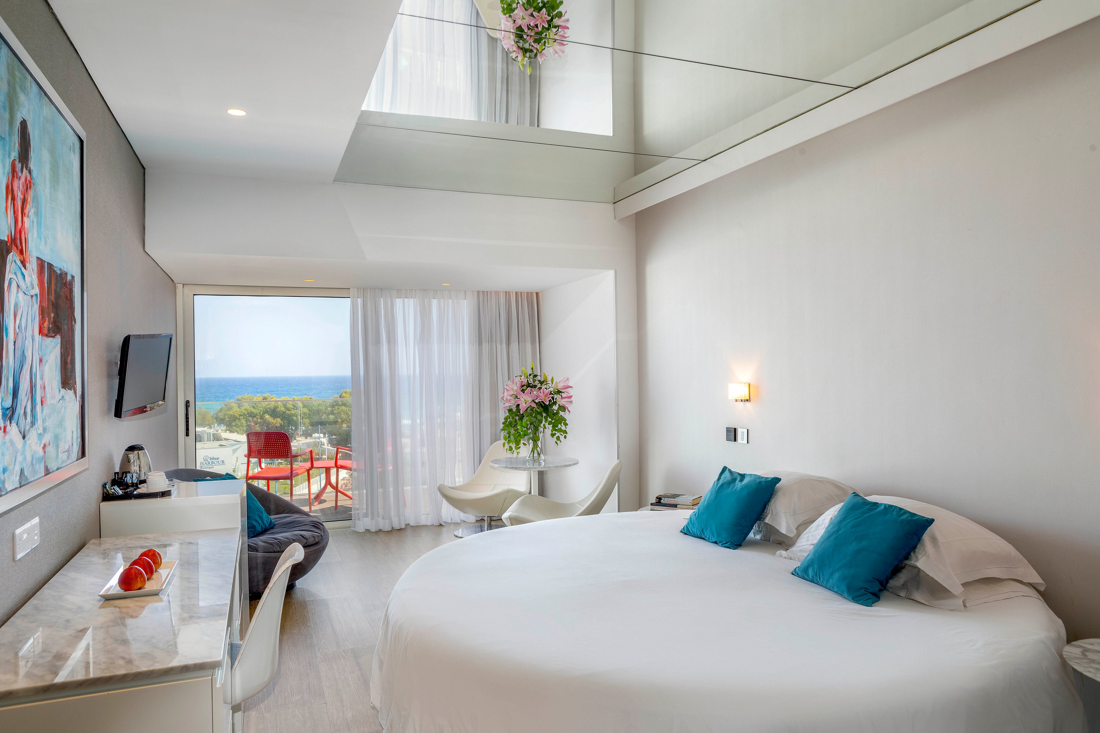 A contemporary hotel room features a large round bed with teal accent pillows and soft white linens. The space is bright, with floor-to-ceiling curtains leading to a balcony that overlooks the ocean and outdoor seating. A desk area, wall-mounted television, and colorful artwork add modern touches, while fresh flowers and soft lighting create a welcoming atmosphere. The overall design is clean, airy, and minimalist, emphasizing comfort and the scenic coastal view.