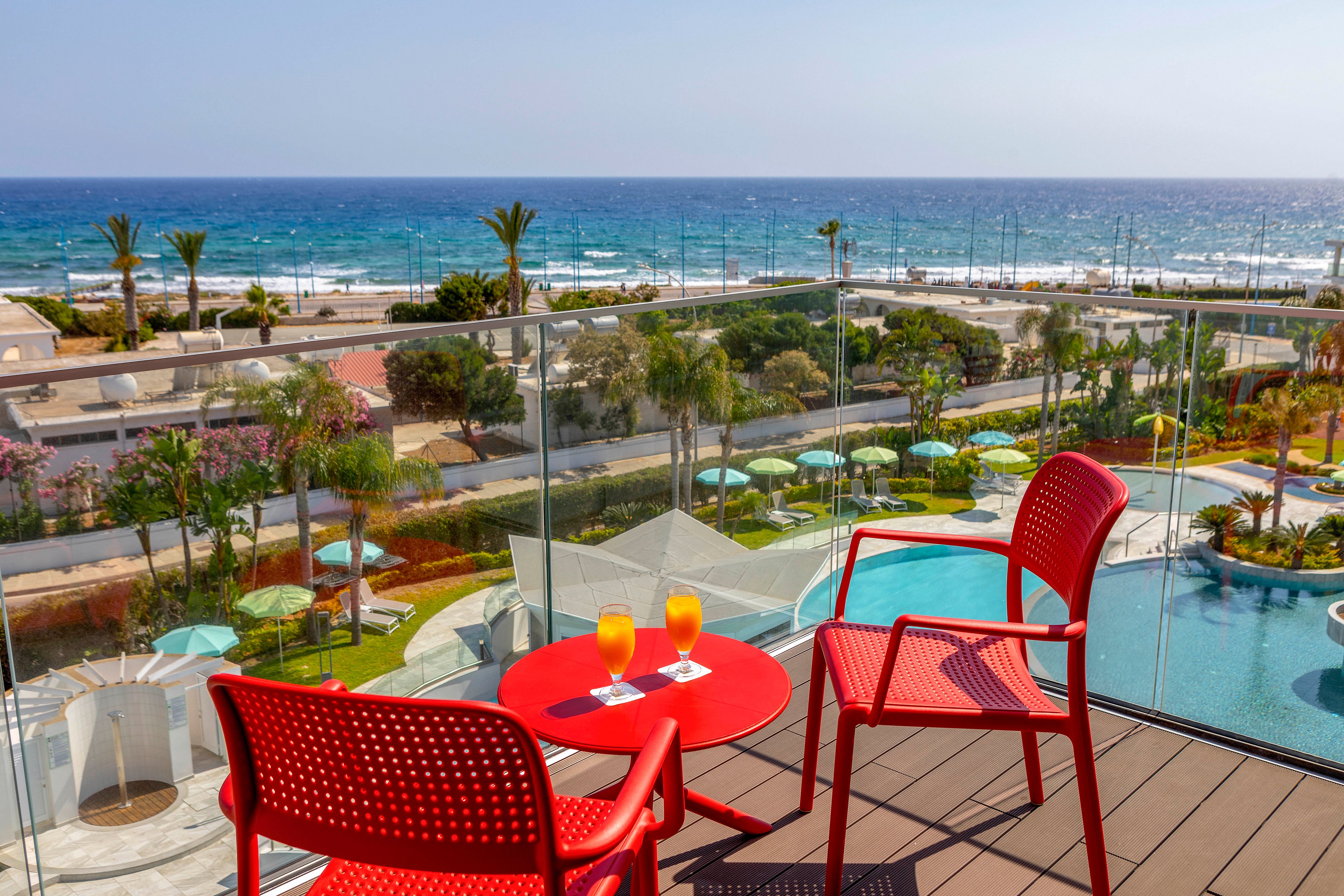 A sunny balcony overlooks a resort pool and a vibrant blue ocean. Two bright red chairs and a matching table hold two orange-colored drinks, suggesting a relaxing vacation moment. Below, palm trees, umbrellas, and landscaped gardens create a tropical atmosphere. The clear sky and sparkling water emphasize a warm, inviting coastal setting.