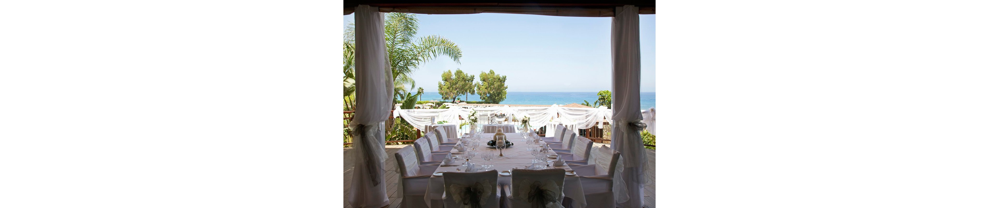 A long, elegantly decorated dining table is set for a formal event on a covered terrace overlooking the sea. White chair covers, tablecloths, and delicate floral arrangements create a sophisticated wedding or celebration atmosphere. Sheer white drapes frame the view of palm trees, greenery, and the blue ocean beyond. Soft natural light enhances the serene and romantic coastal setting.