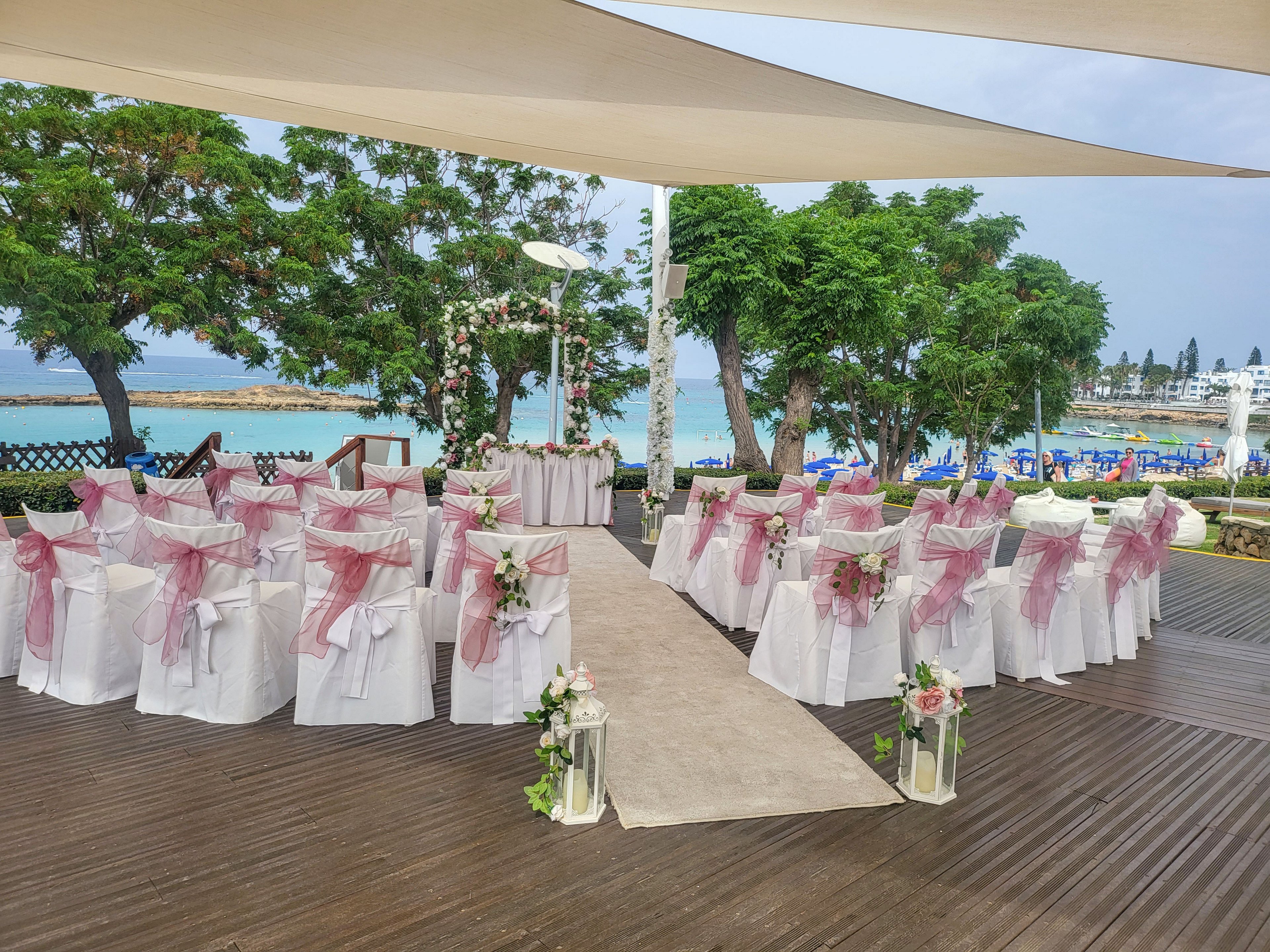 An elegant outdoor wedding ceremony is arranged on a wooden deck overlooking the sea. White chairs decorated with pink organza bows are aligned on both sides of a central aisle leading to a floral arch and altar table. Green trees frame the venue while a beach and blue water are visible in the background. Soft pastel decorations and fresh flowers create a romantic, coastal atmosphere.