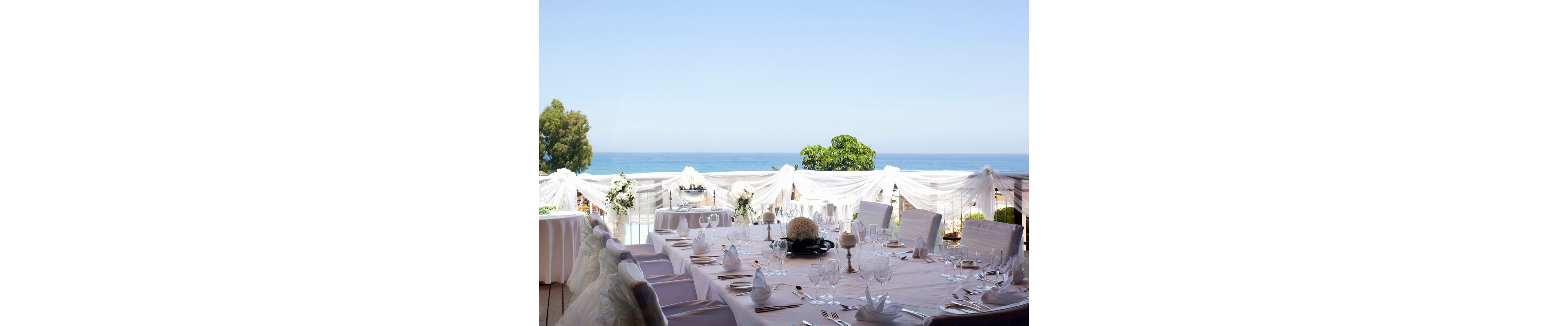 A large round table is elegantly set for a formal event on a terrace overlooking the sea. White linens, folded napkins, and neatly arranged glassware create a refined and sophisticated atmosphere. Sheer white drapes and floral arrangements decorate the railing, framing the ocean view in the background. The bright daylight and soft pastel tones contribute to a serene, romantic mood suitable for a wedding or special celebration.