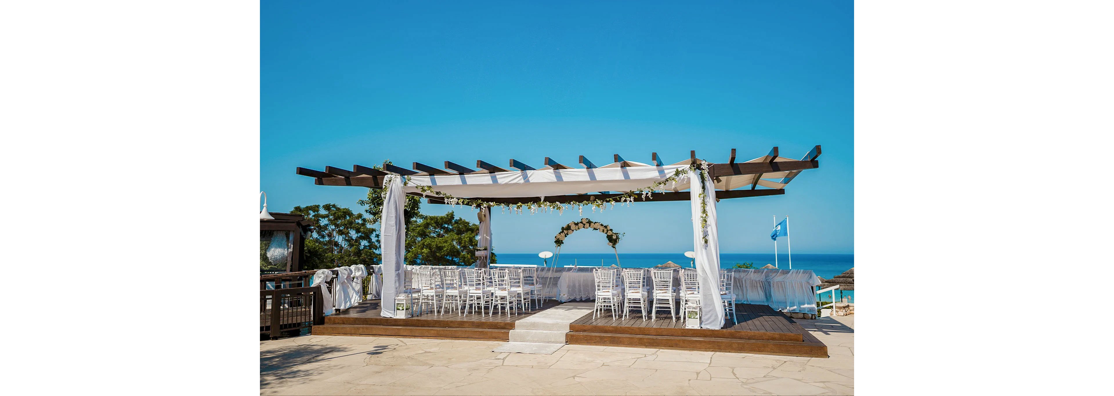 An elegant outdoor wedding ceremony area is arranged beneath a wooden pergola draped with white fabric. Rows of white chairs face a floral arch at the front, creating a formal seating arrangement. The venue is set on a raised wooden platform overlooking the sea under a clear blue sky. Greenery and floral garlands decorate the structure, adding a romantic touch to the coastal setting.