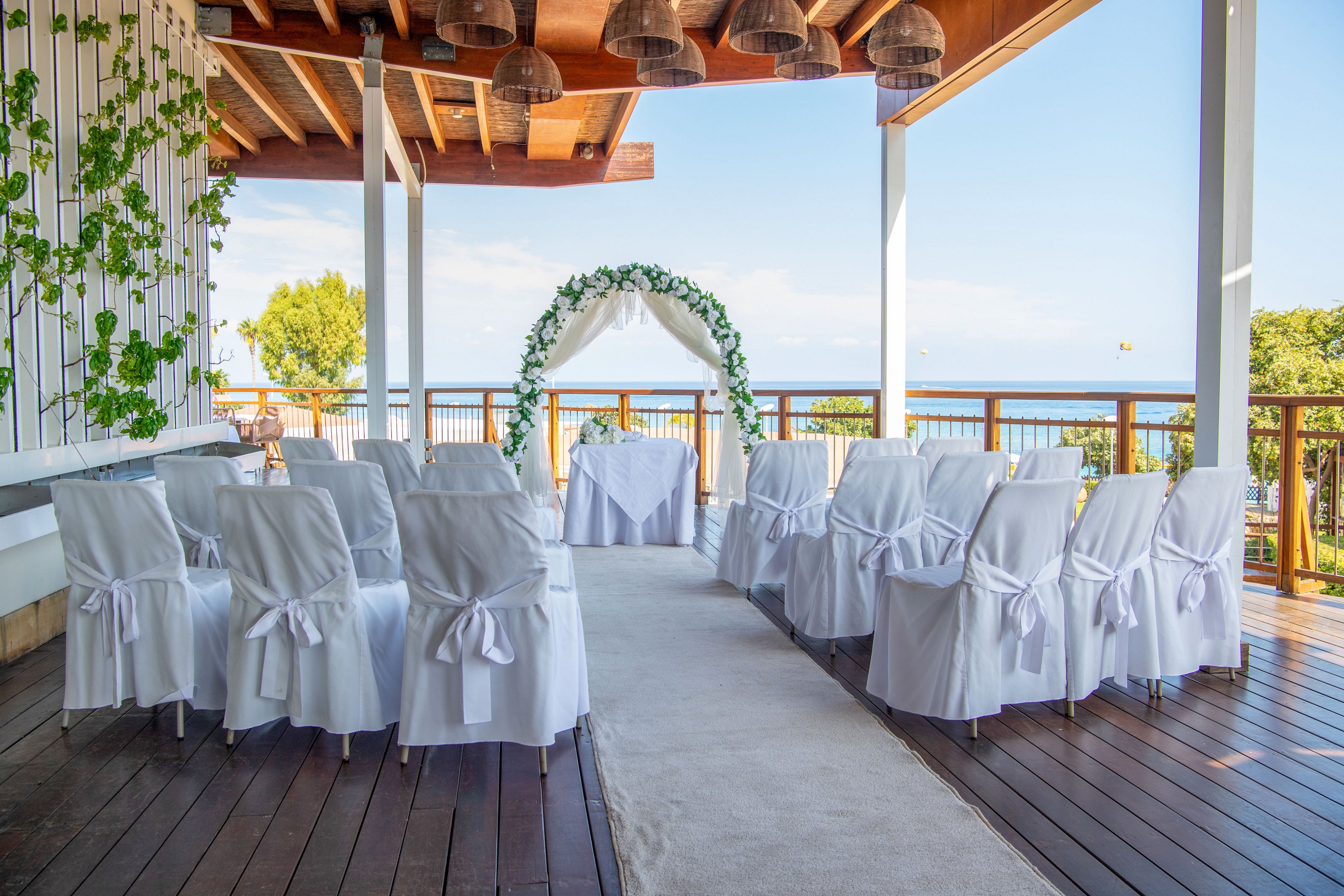 An elegant wedding ceremony setup is arranged on a covered wooden terrace overlooking the sea. White chairs with satin bows face a floral arch and a small table at the front. The open-air venue features wooden beams, hanging lights, and greenery climbing up one side. Bright daylight and ocean views create a romantic and serene atmosphere.