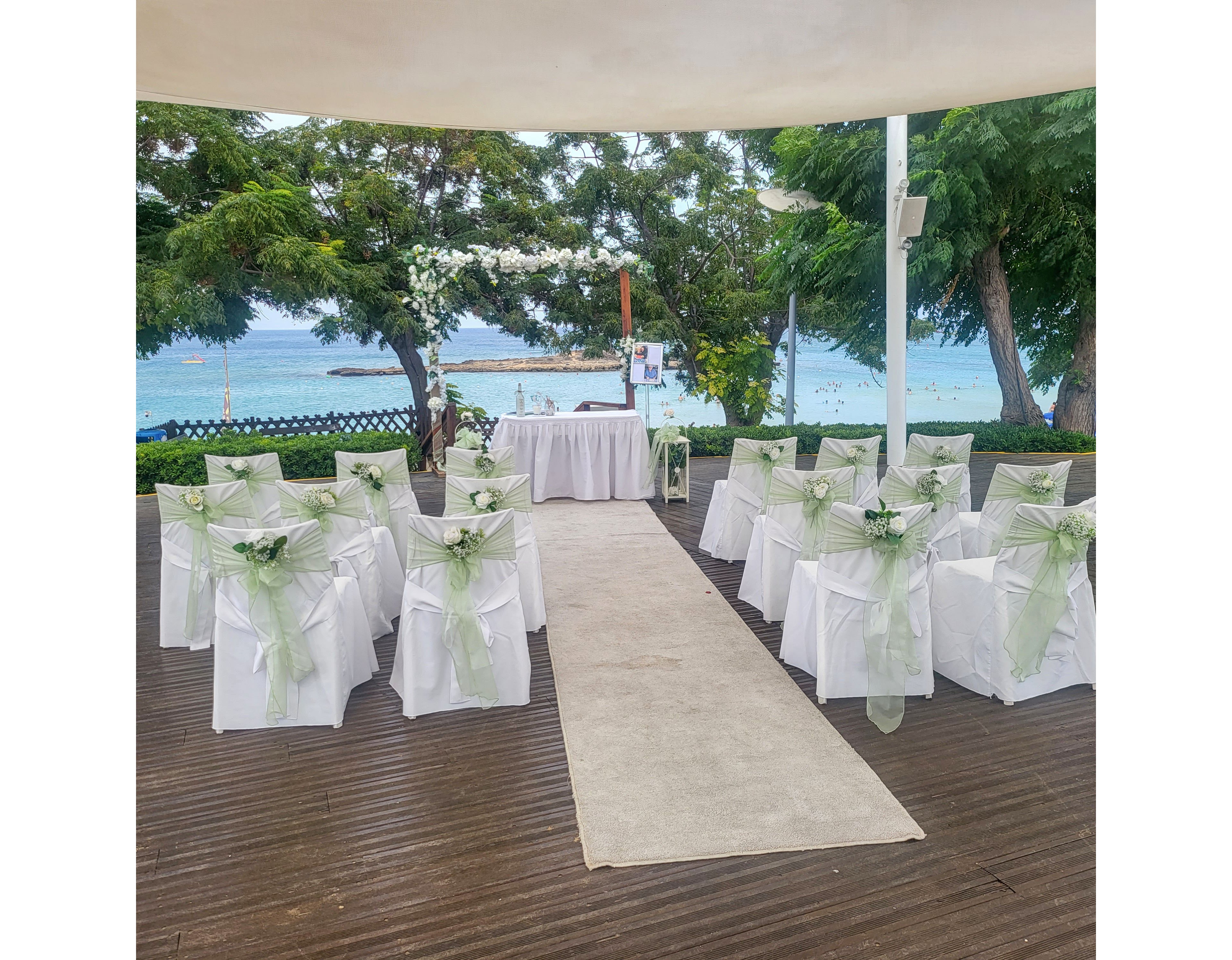 An elegant outdoor wedding ceremony setup overlooks a calm seaside backdrop. White chairs decorated with soft green ribbons and floral arrangements line both sides of a central aisle. At the end of the aisle, a small table and archway adorned with flowers frame the view of the ocean. Lush green trees surround the venue, creating a serene and intimate atmosphere. The overall scene suggests a romantic coastal celebration ready for guests to arrive.