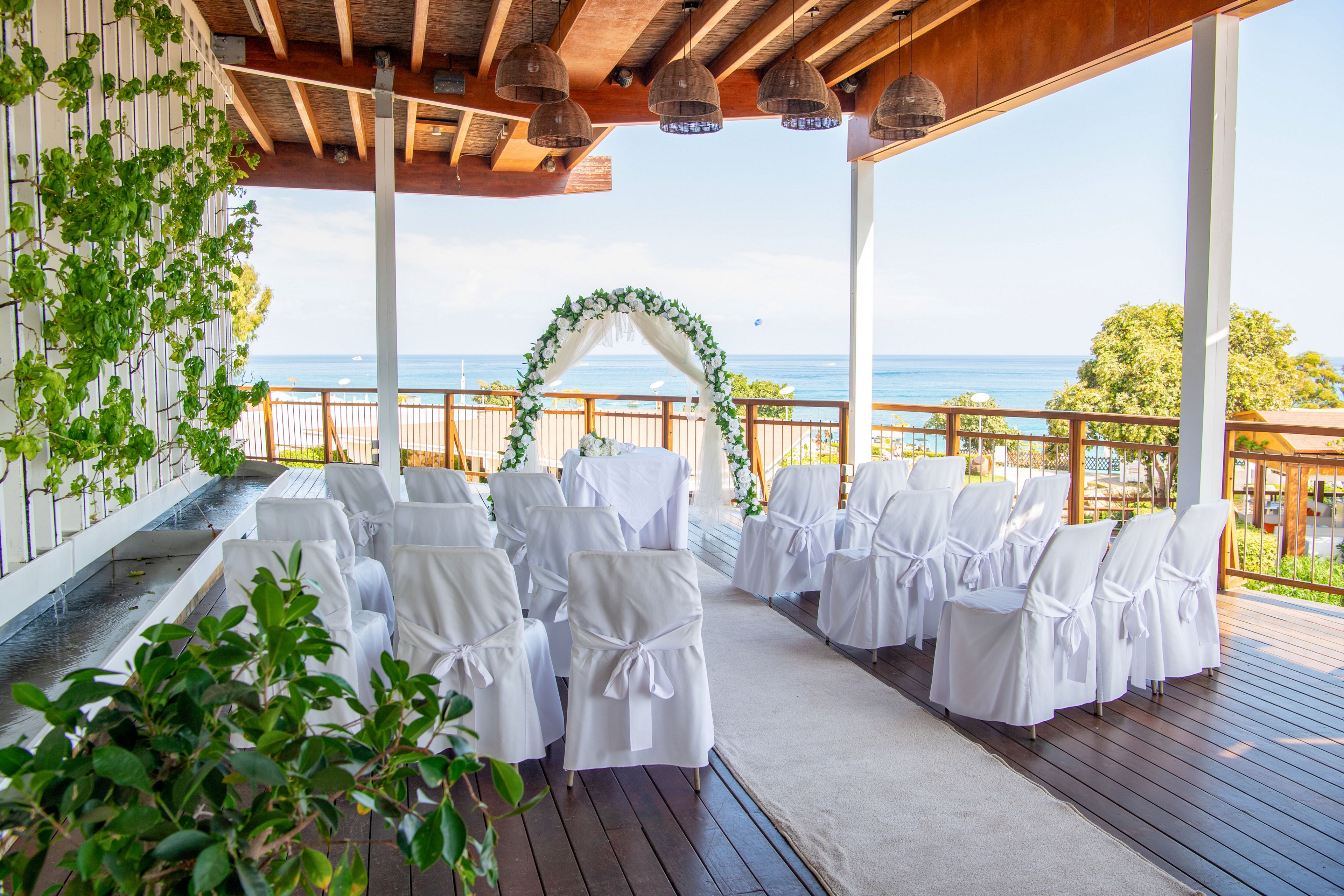 An outdoor wedding ceremony area is arranged on a wooden terrace overlooking the sea. White chairs with bows are lined in rows facing a floral arch at the end of the aisle. Green plants climb the wall and frame the space, adding a natural touch. The bright daylight and ocean view create a serene and romantic atmosphere.