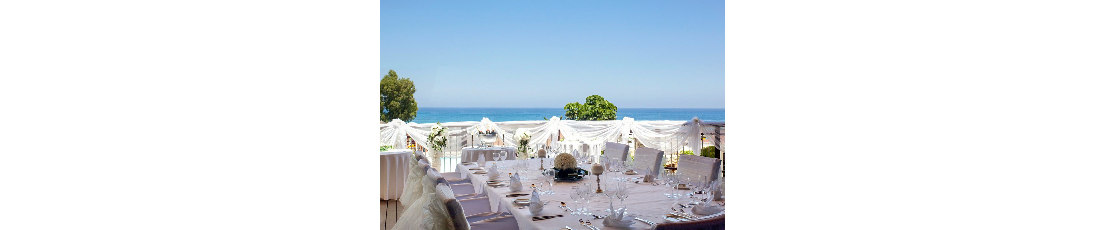 A large round table is elegantly set for a formal event overlooking a calm blue sea. White tablecloths, folded napkins, glassware, and plates are neatly arranged, suggesting a wedding reception or special celebration. Sheer white drapes and floral arrangements decorate the balcony railing, framing the ocean view. The bright daylight and clear sky create a serene and romantic atmosphere.