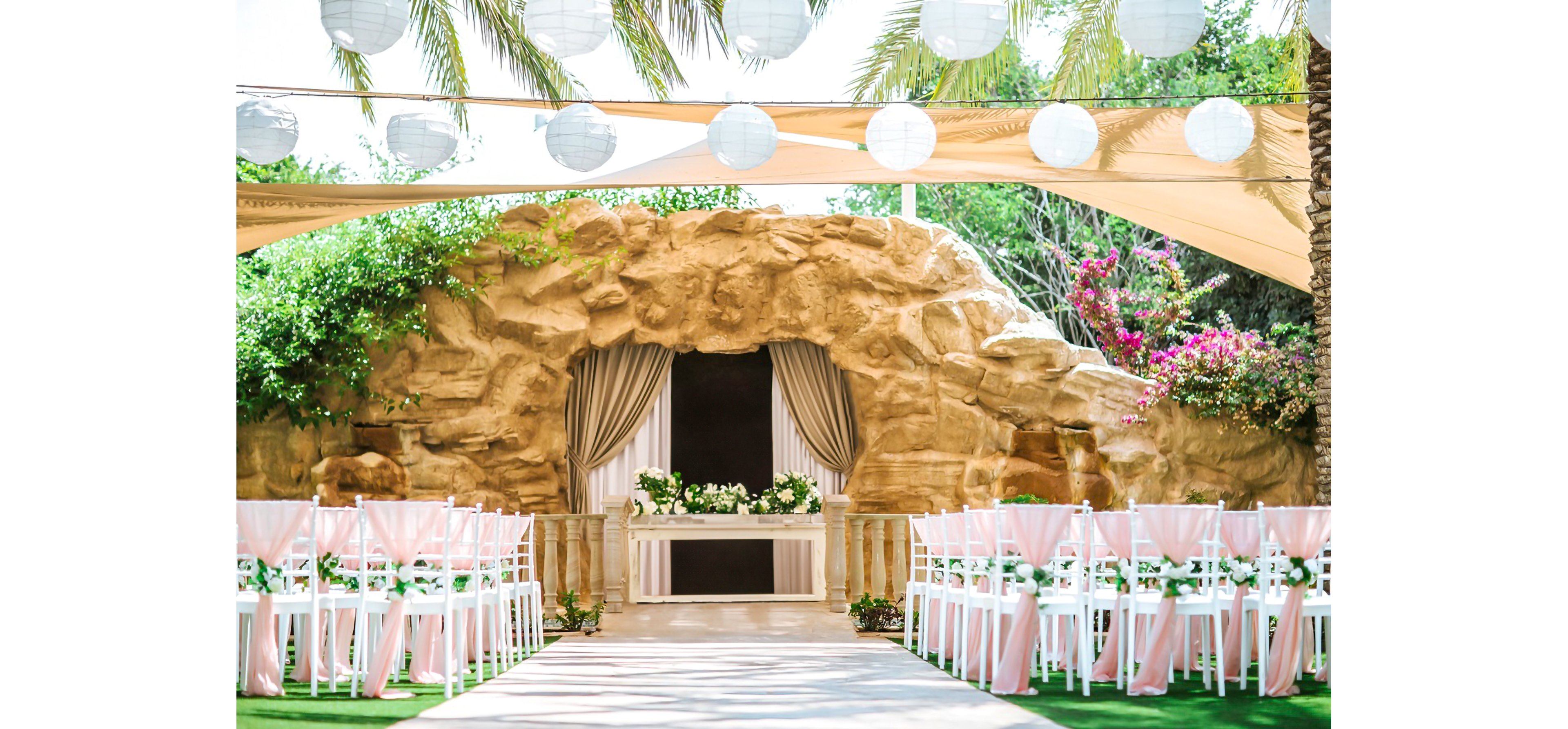 An elegant outdoor wedding ceremony setup features rows of white chairs with soft pink fabric lining a central aisle. The aisle leads to a rocky grotto-style backdrop with draped curtains and a flower-adorned table at the front. Overhead, round white paper lanterns hang beneath shade sails, while palm trees and lush greenery frame the scene. Bright natural light and pastel colors create a romantic, festive atmosphere.