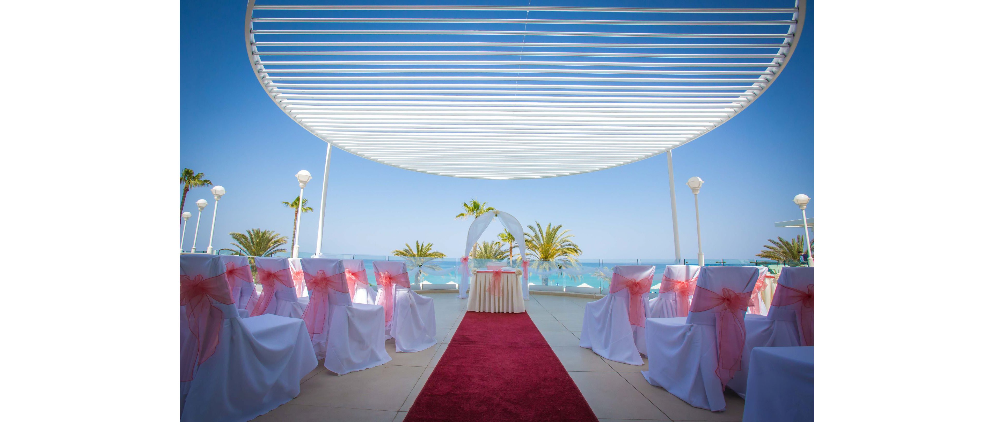 An elegant outdoor wedding ceremony setup overlooks a bright blue ocean. White chairs decorated with pink organza bows line both sides of a red carpet aisle. A small table and white arch stand at the end of the aisle under a modern open-air canopy. Palm trees and clear skies create a tropical, romantic atmosphere. The scene is calm, festive, and ready for guests to arrive.