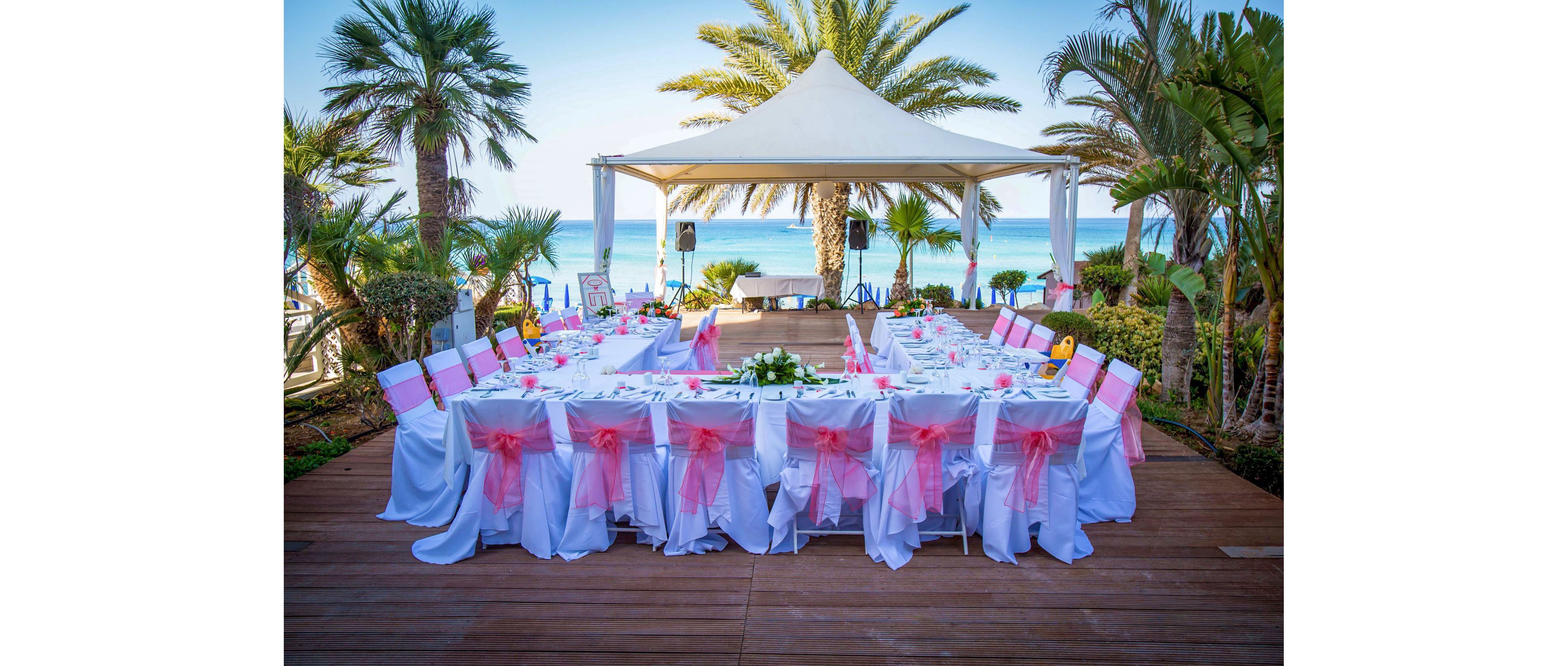 An elegant outdoor reception setup is arranged beneath a white canopy overlooking a tropical beach. Long banquet tables are covered with white linens and decorated with pink chair sashes and floral centerpieces. Palm trees surround the wooden deck, framing the view of the turquoise ocean in the background. The scene suggests a festive wedding or special event in a coastal resort setting.