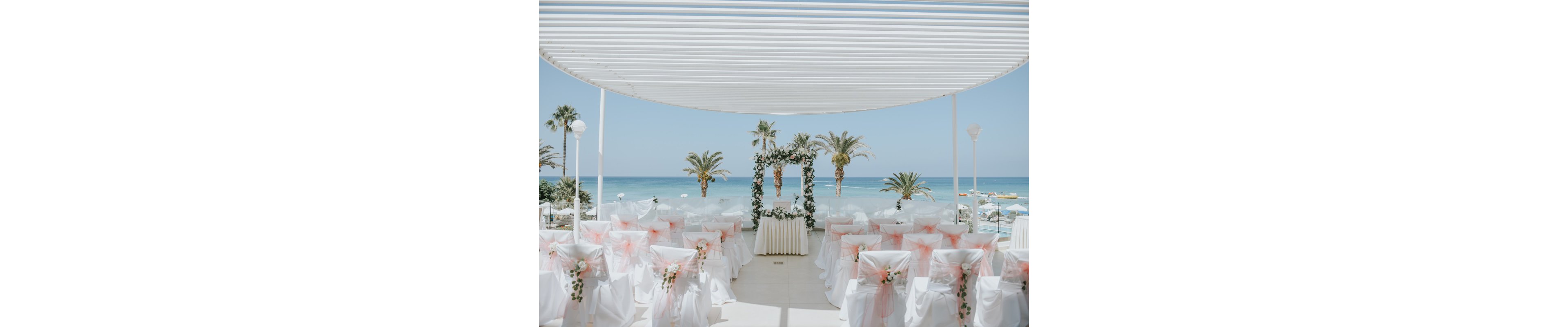 An outdoor wedding ceremony space is arranged under a modern white pergola overlooking a bright blue sea. Rows of white chairs are decorated with soft pink tulle bows and floral arrangements, facing a floral arch at the front. Palm trees and calm ocean water create a tropical backdrop, with boats visible in the distance. The overall scene feels airy, romantic, and ready for guests to arrive.
