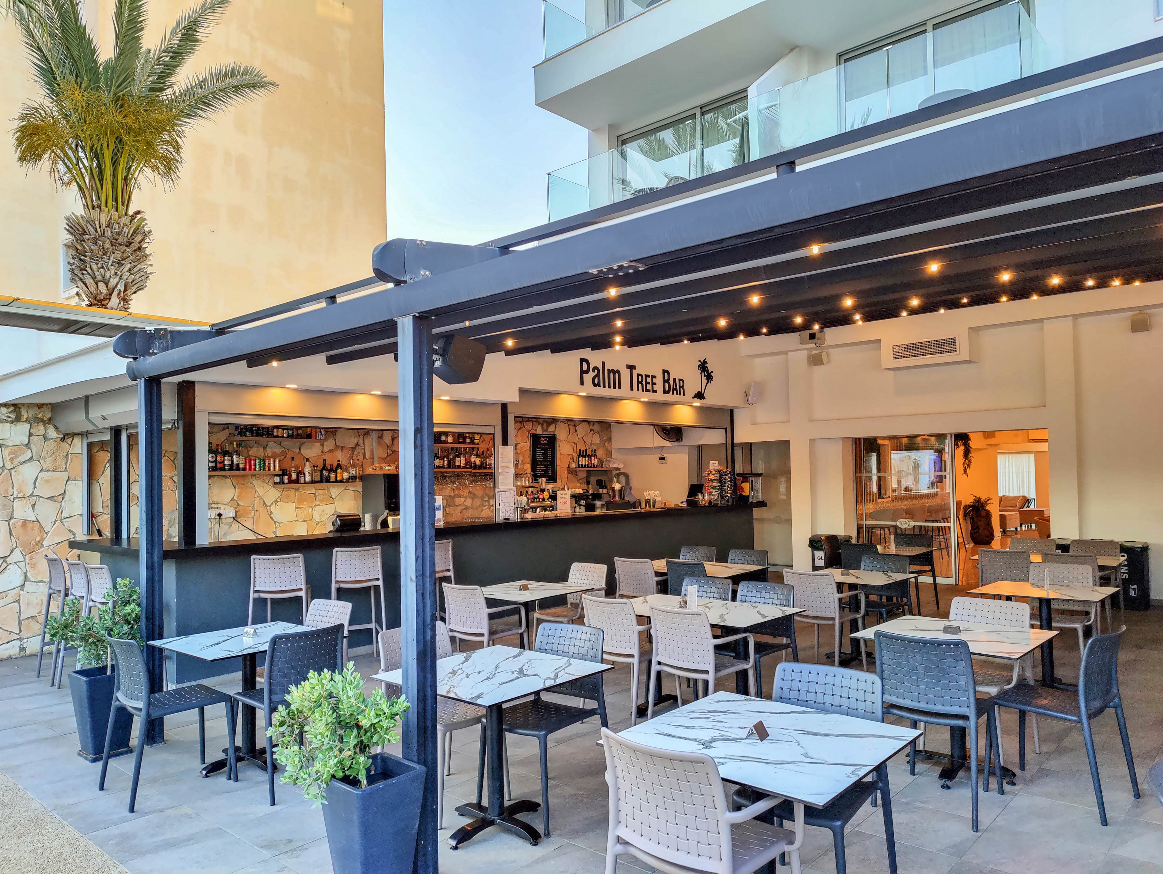 Open-air bar named Palm Tree Bar with modern terrace seating is shown in a sunny resort setting. Multiple tables and chairs are arranged neatly under a pergola with string lights, creating a relaxed atmosphere. The bar counter displays bottles and glassware, with potted plants adding greenery around the seating area. A contemporary building facade and palm tree in the background suggest a vacation or leisure environment.