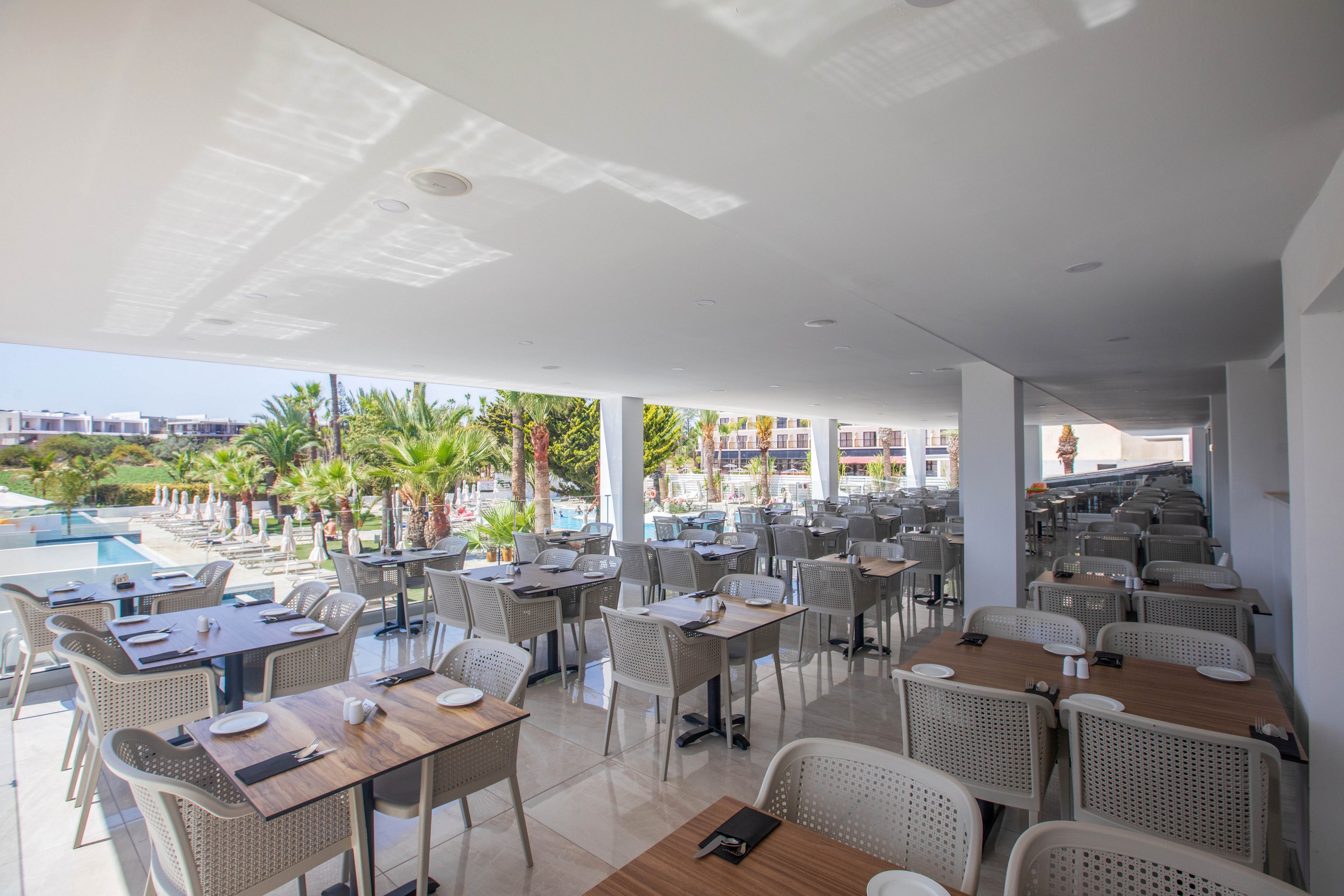 Spacious open-air restaurant seating area overlooks a resort swimming pool lined with sun loungers and palm trees. Numerous tables and light-colored chairs are neatly arranged under a shaded terrace, prepared with placemats and cutlery. Bright daylight reflects off the white ceiling, creating a clean and modern atmosphere. In the background, resort buildings frame the relaxing holiday setting.