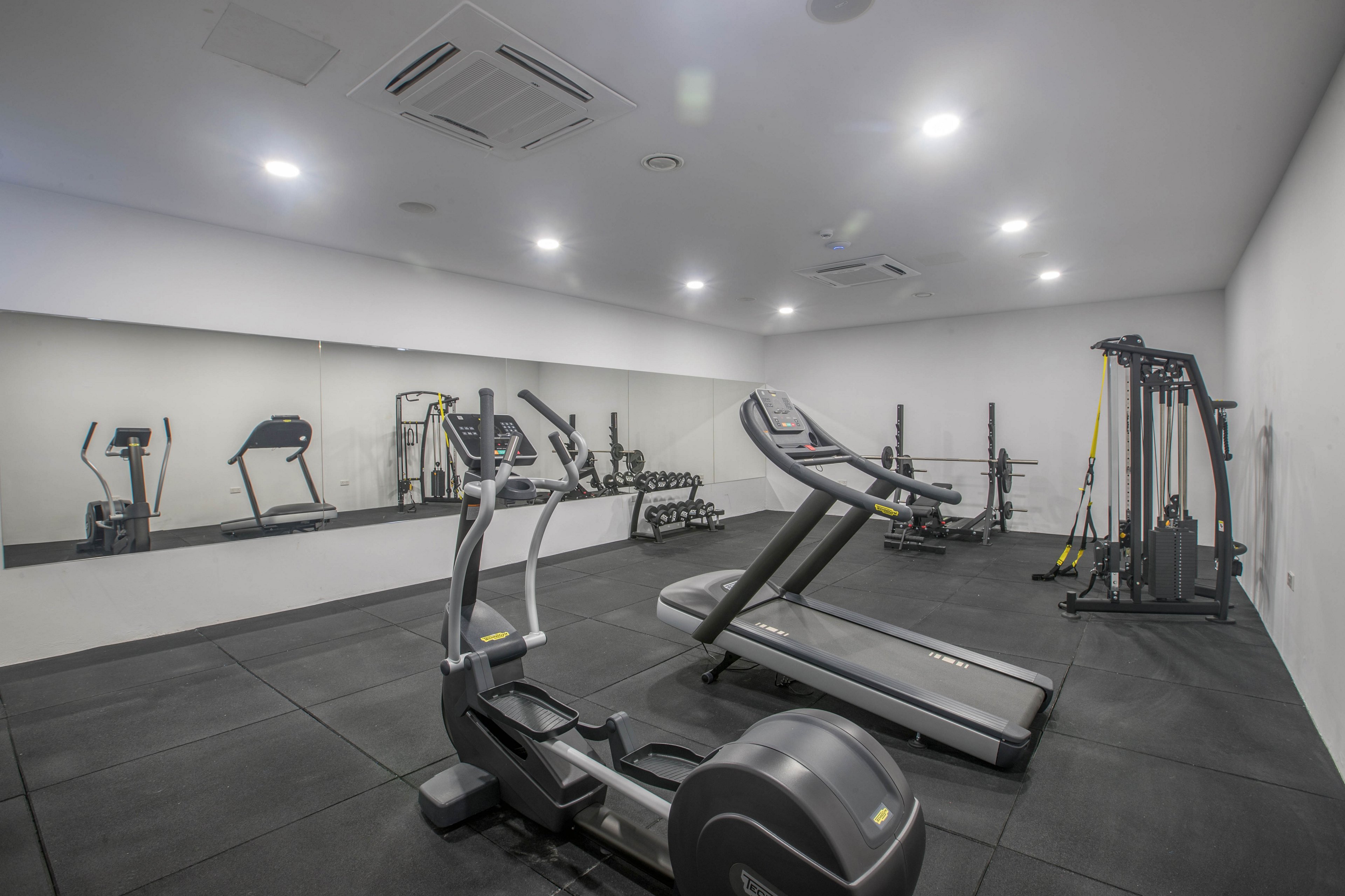 A clean, modern indoor gym features a treadmill and elliptical trainer in the foreground. In the background, weight benches, dumbbells, and strength machines are arranged along a mirrored wall. The room has bright overhead lighting, neutral gray flooring, and white walls that create a minimalist atmosphere. The space appears organized and ready for personal fitness workouts.