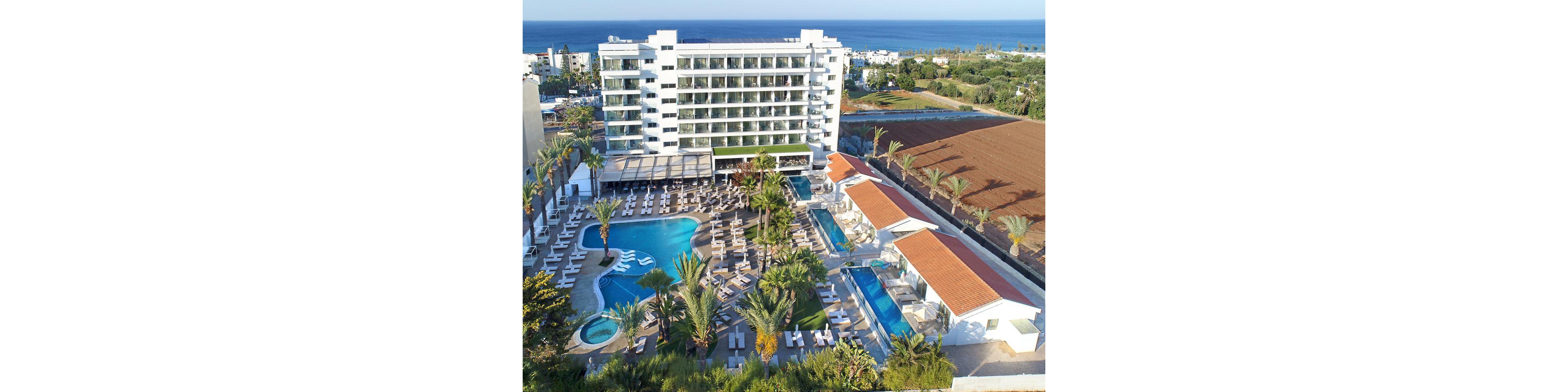 A large multi-story resort hotel is shown from above, featuring several outdoor swimming pools surrounded by sun loungers and palm trees. The complex includes a main curved pool, smaller rectangular pools, and white cabanas with red-tiled roofs. The setting appears coastal, with the sea visible in the background and nearby residential buildings and fields. The overall scene suggests a sunny vacation destination focused on relaxation and leisure by the water.