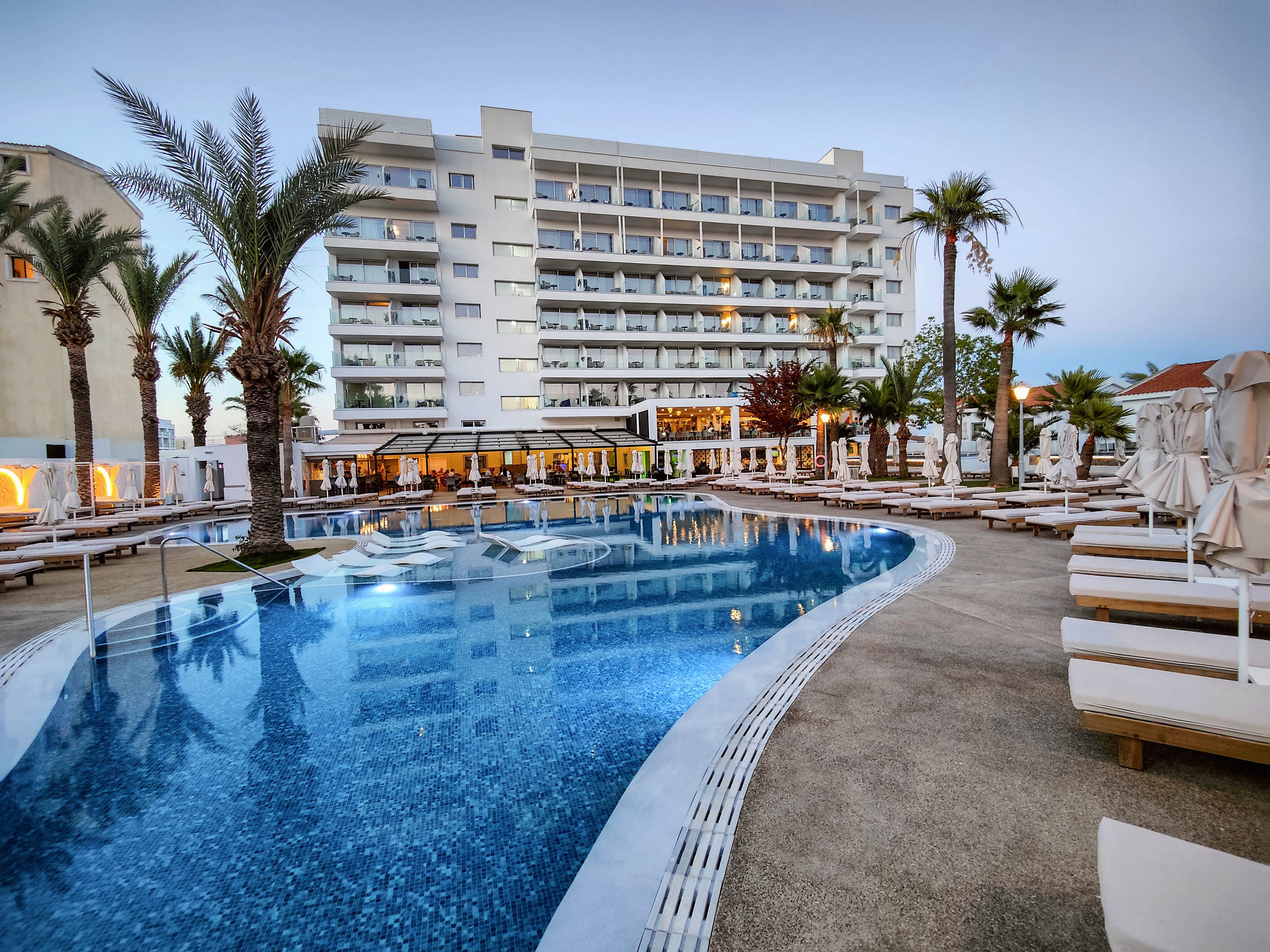 A large curved swimming pool is surrounded by white sun loungers and umbrellas at a modern resort. A multi-story hotel building rises in the background, with balconies overlooking the pool area. Palm trees and soft evening lighting create a tranquil vacation atmosphere. The sky is clear and blue, suggesting a warm coastal or tropical setting.