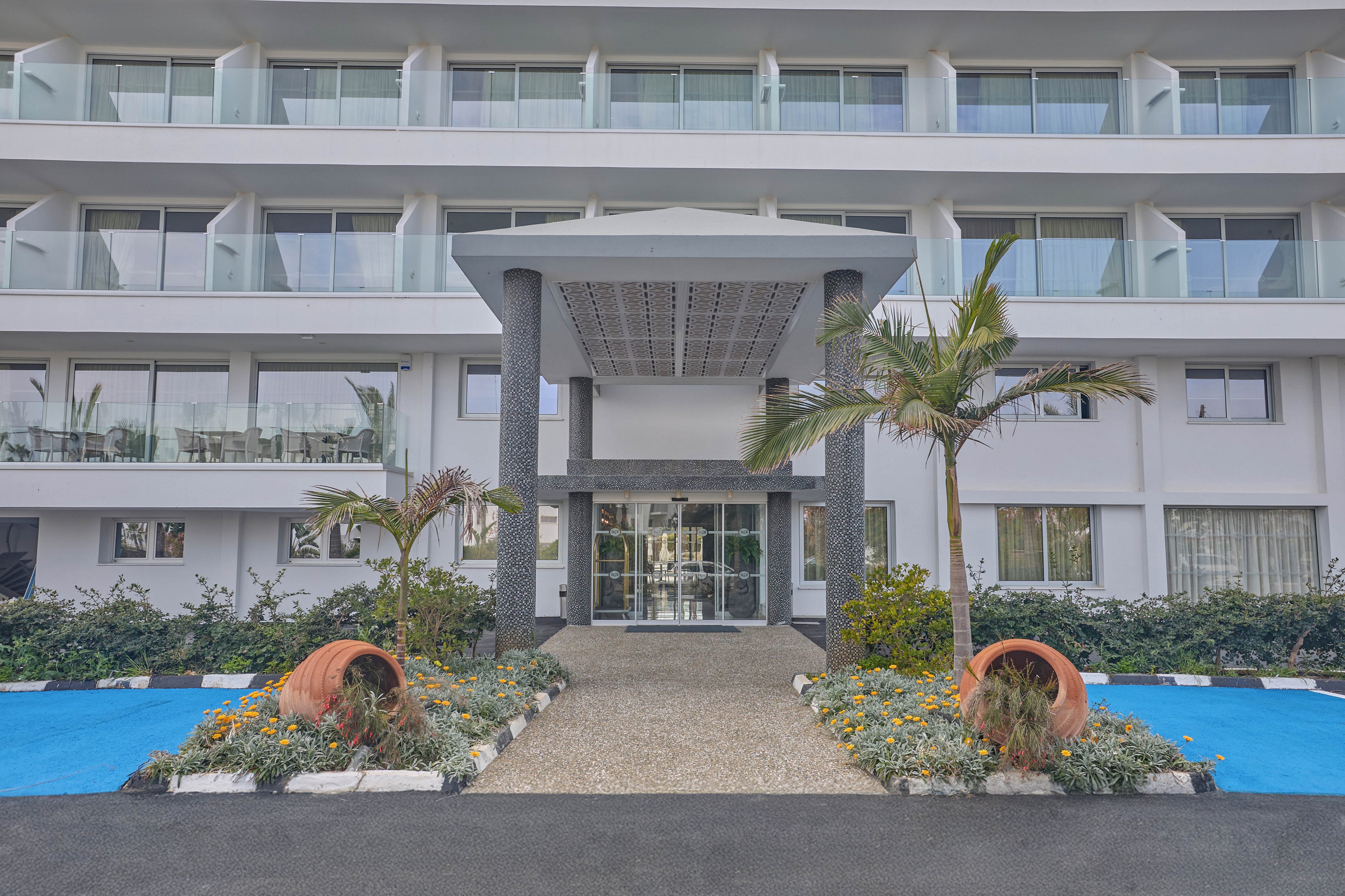A contemporary multi-story hotel building features a central glass entrance framed by tall columns. The foreground shows a landscaped garden with palm trees, flowering plants, and large terracotta pots on either side of the walkway. Blue decorative ground surfaces contrast with the greenery and the neutral tones of the facade. Balconies line the upper floors, suggesting guest rooms overlooking the entrance area.