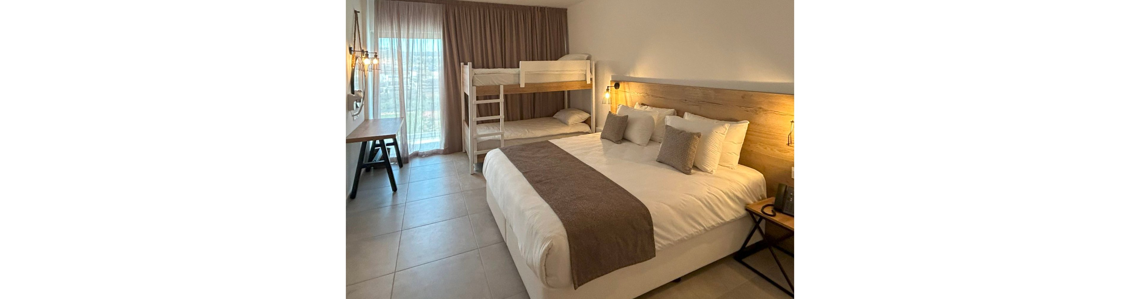 A contemporary hotel room features a large double bed with white linens and a taupe runner. In the corner, a white bunk bed provides additional sleeping space, suggesting family-friendly accommodation. Soft neutral tones, wood accents, and tiled flooring create a clean, minimalist aesthetic. A desk area, bedside lamps, and sheer curtains over a large window complete the cozy, functional space.