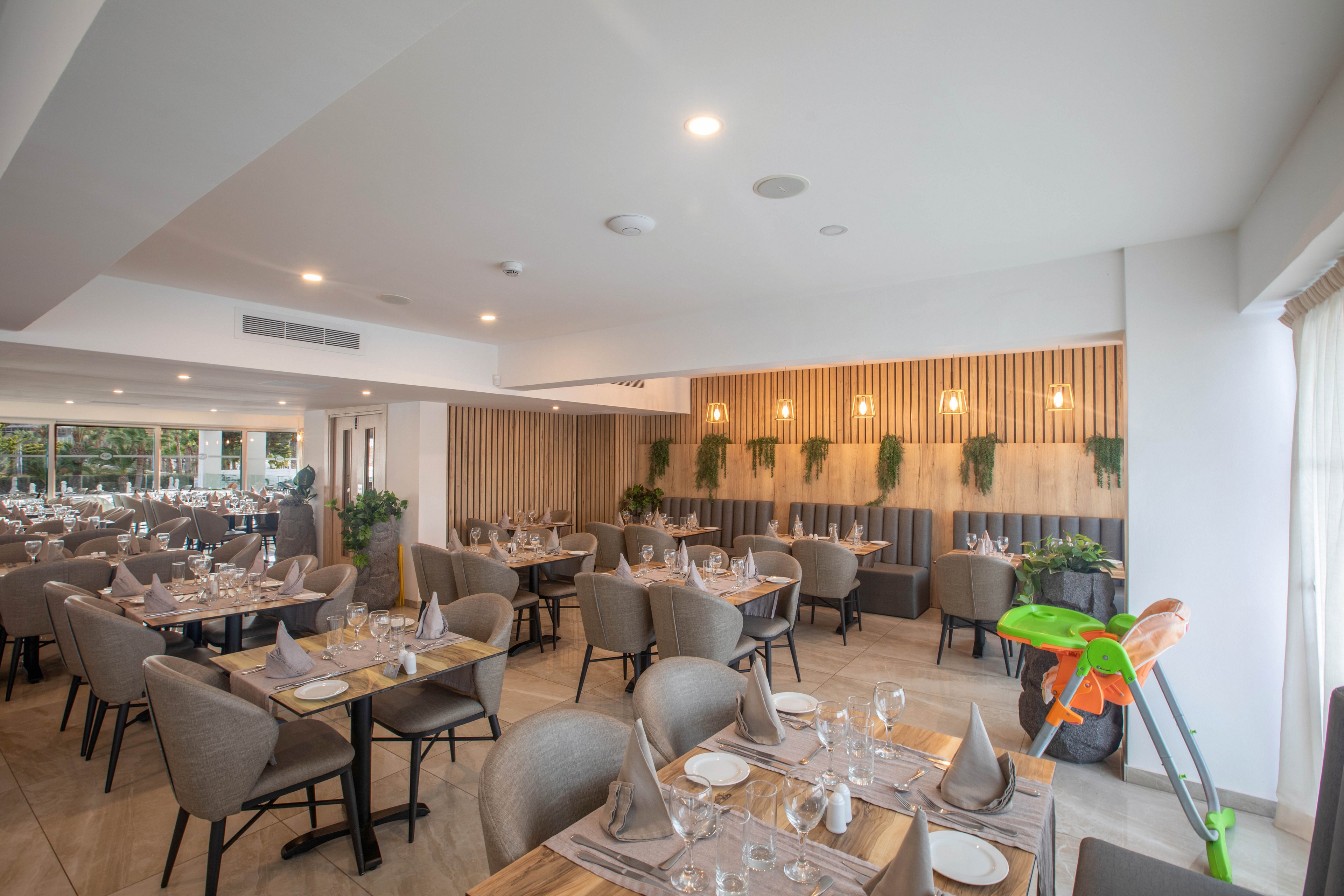 A contemporary restaurant dining room is neatly arranged with multiple tables set for guests. Neutral-toned upholstered chairs and wooden tables create a warm, inviting atmosphere. Each table is prepared with folded napkins, plates, cutlery, and glassware, ready for service. Soft lighting, vertical wooden slats, and green plants decorate the back wall, while large windows bring in natural light and a small children's high chair adds a family-friendly touch.