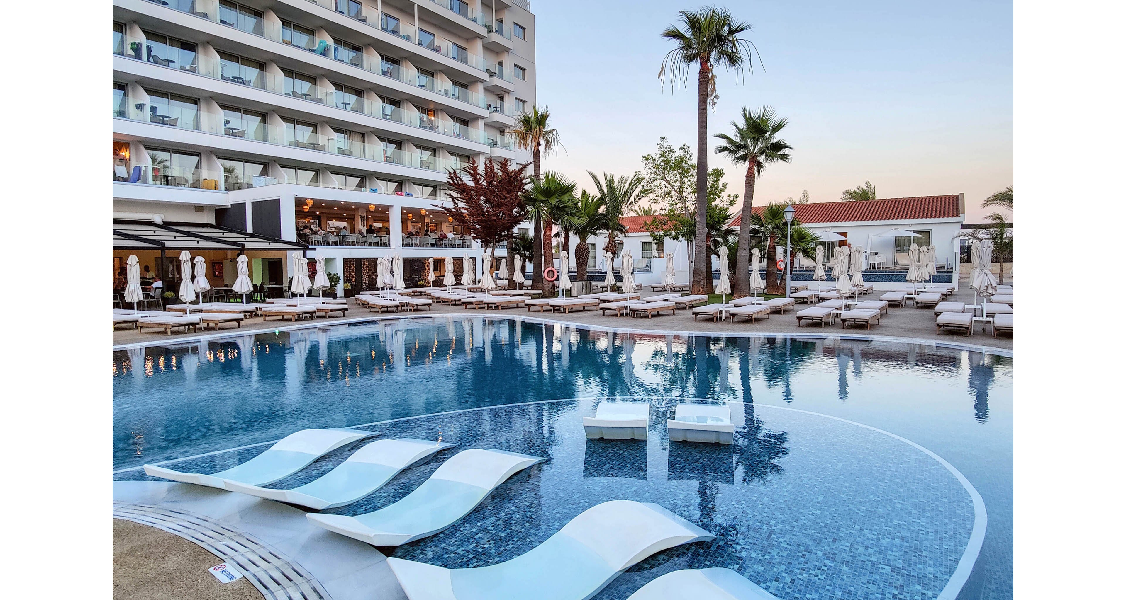 A contemporary multi-story resort hotel overlooks a large curved swimming pool with in-water loungers. Numerous sunbeds and umbrellas line the pool deck, creating a relaxed vacation atmosphere. Palm trees and landscaped greenery add a tropical touch, while soft evening light enhances the tranquil setting. The scene suggests a stylish, upscale destination for leisure and relaxation.