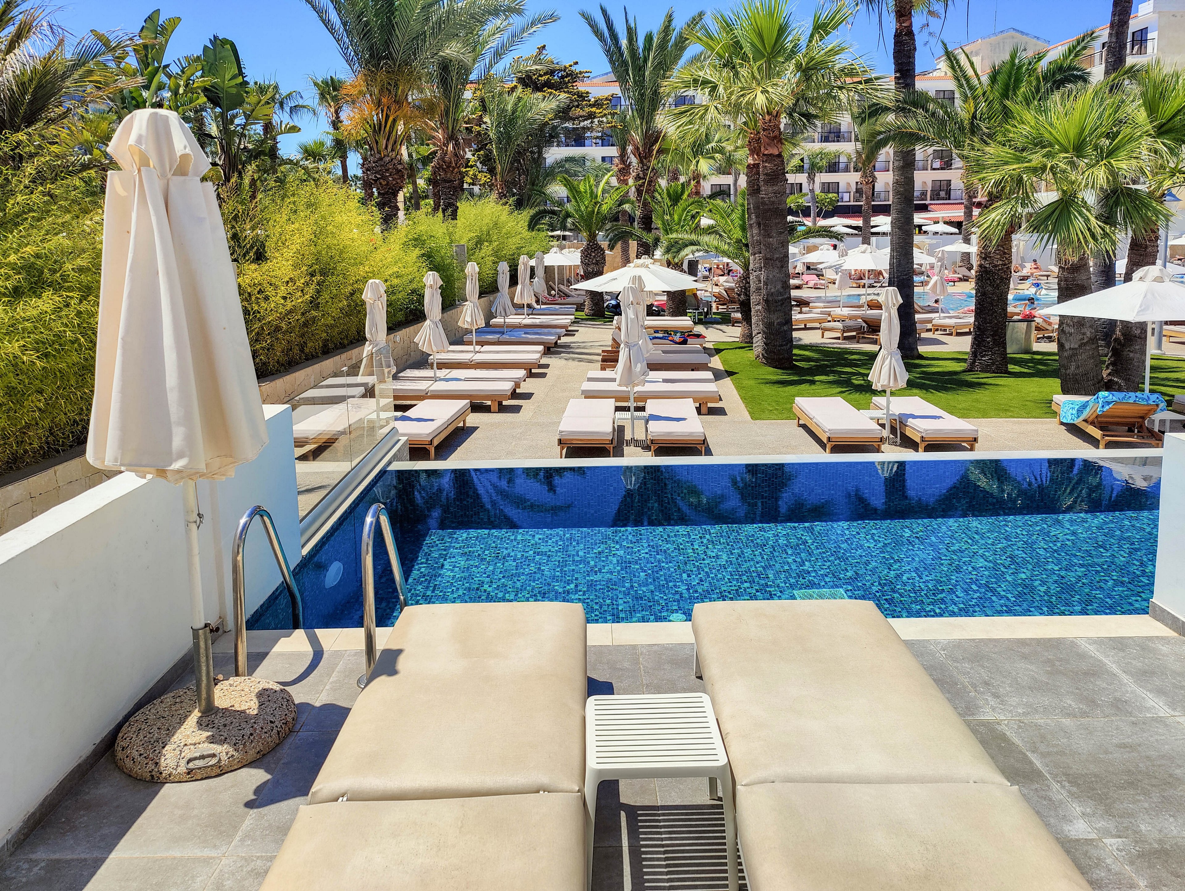 Image of a luxury resort pool area with two sun loungers in the foreground beside a small infinity-style swimming pool. Beyond the pool, multiple cushioned loungers with umbrellas are arranged neatly on a lawn lined with tall palm trees. In the background, additional seating, umbrellas, and resort buildings suggest a busy holiday destination near the water. The scene is bright and sunny, emphasizing relaxation and vacation leisure.