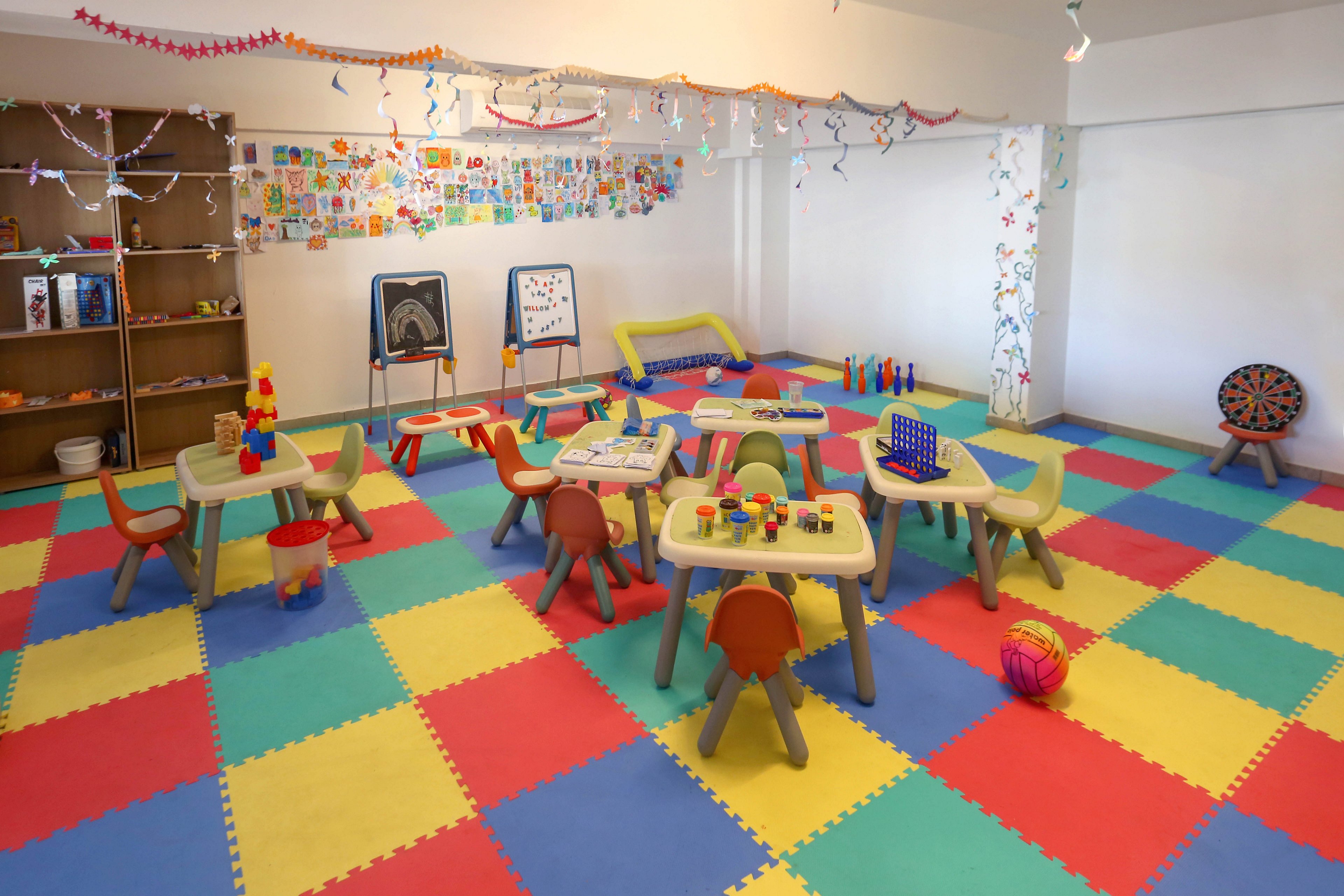 Brightly colored preschool classroom with small tables and chairs arranged on a multicolor foam tile floor. Various educational toys, blocks, and games are placed on the tables, inviting hands-on play and learning. The walls are decorated with children’s artwork, garlands, and playful patterns, creating a cheerful atmosphere. Shelving and soft play areas in the background suggest a space designed for early childhood education and creative activities.