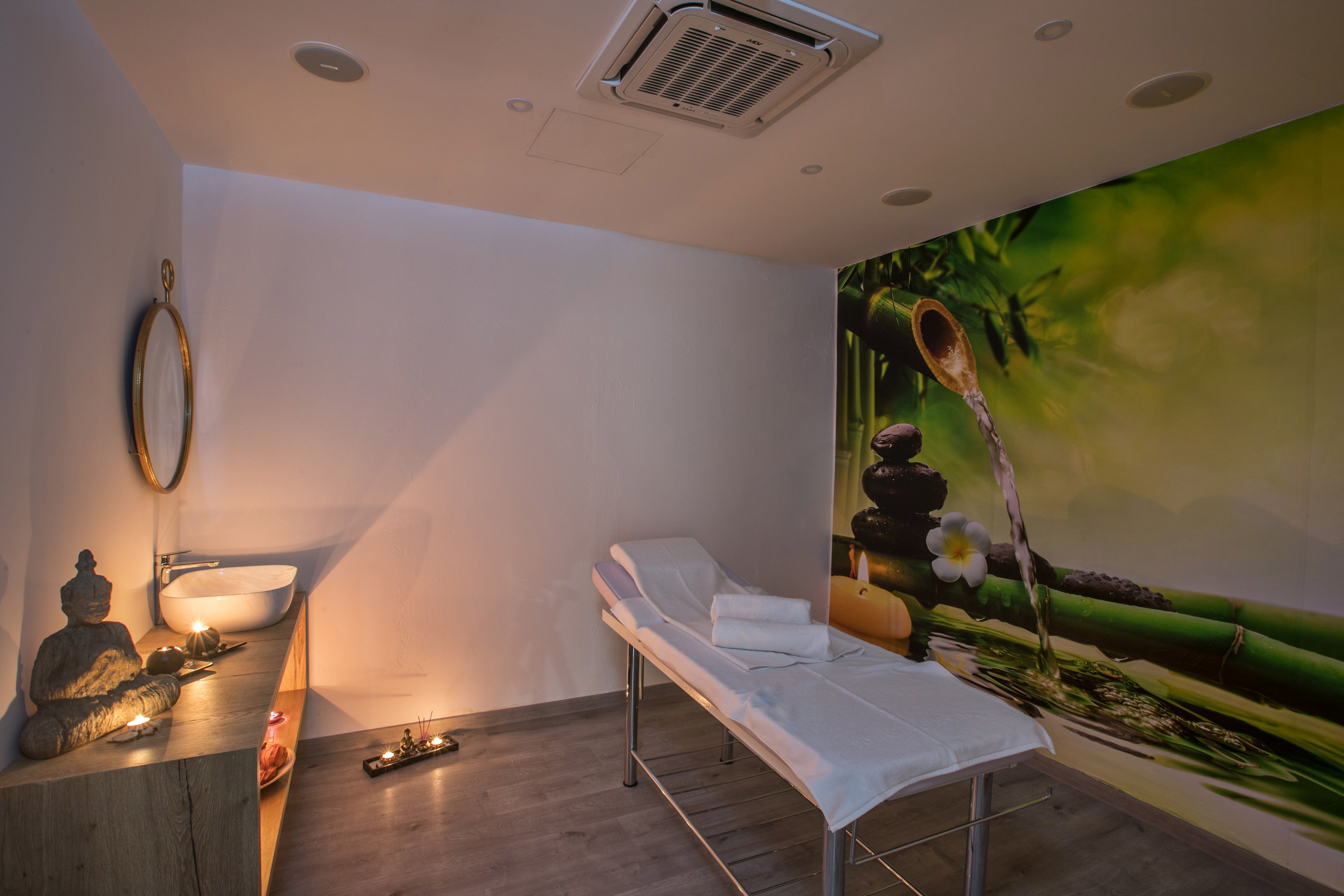 A tranquil spa treatment room features a massage table covered with white linens and neatly folded towels. Soft, warm lighting and candles create a relaxing atmosphere along the wall and wooden cabinet. A decorative Buddha statue and round mirror enhance the peaceful, minimalist decor. The large wall mural shows bamboo, flowing water, and flowers, reinforcing the calm, nature-inspired setting.